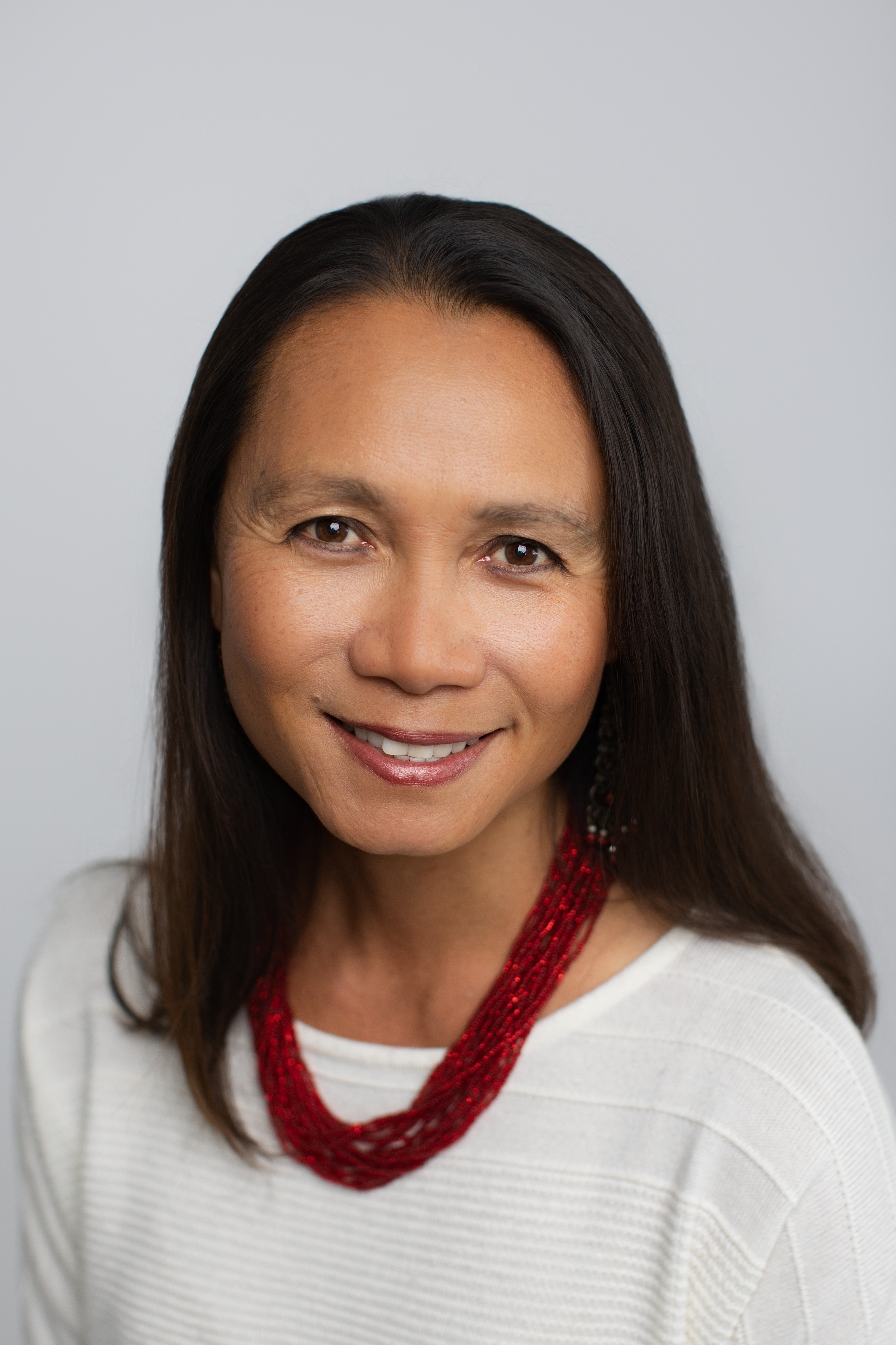 Woman with long dark hair smiling, wearing a white top and red necklace, against a light gray background.