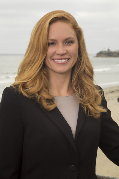 Woman with long wavy hair smiling at the beach, wearing a black blazer and light top, ocean and shoreline in background.