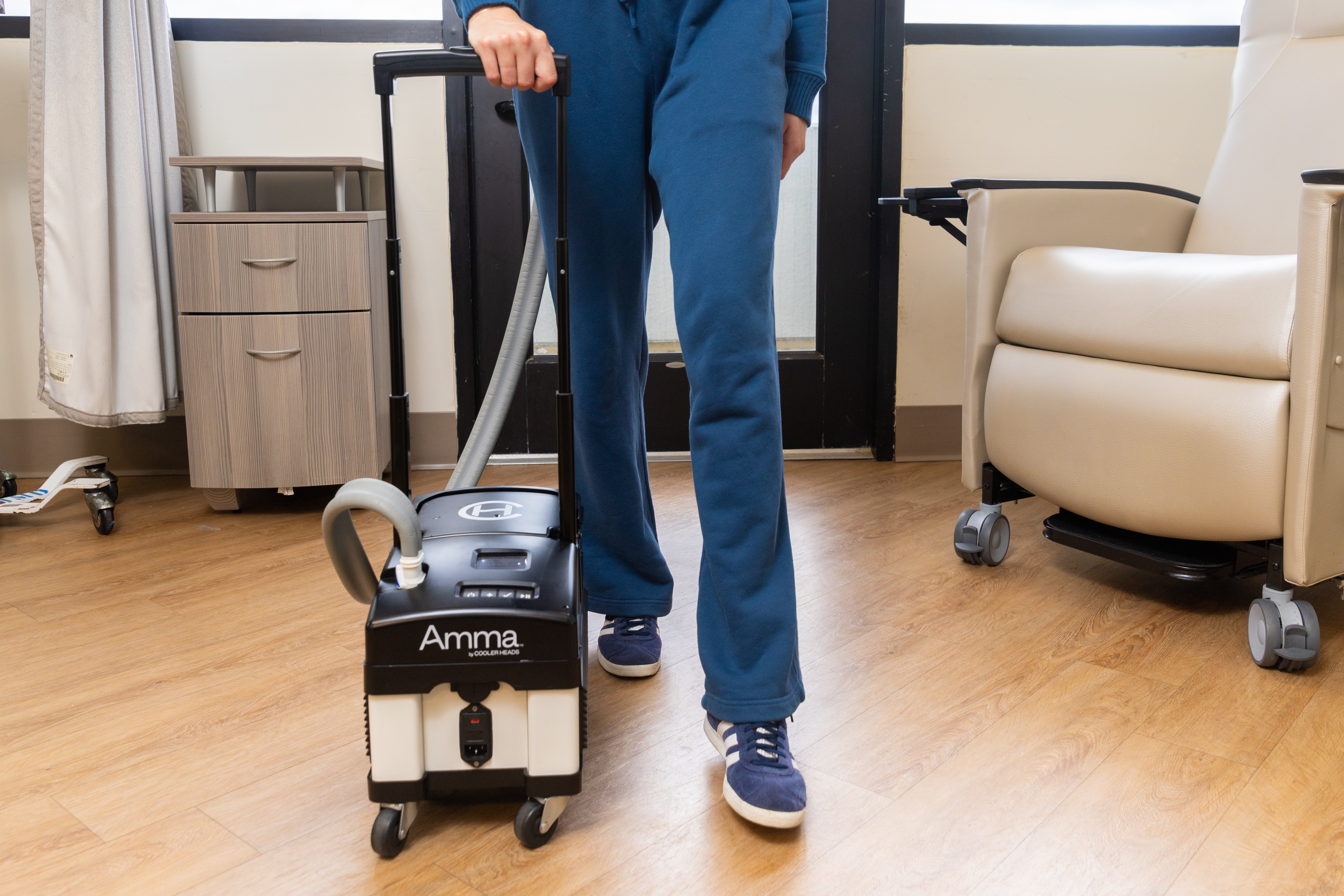 Person standing with a vacuum cleaner in a room with a chair and cabinet, hardwood floor, and large window.