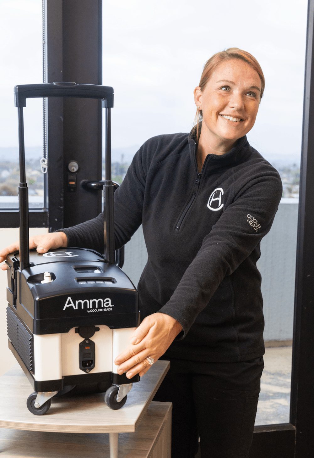 Smiling woman in black jacket with logo, standing next to medical device on table, in room with large windows, city view.