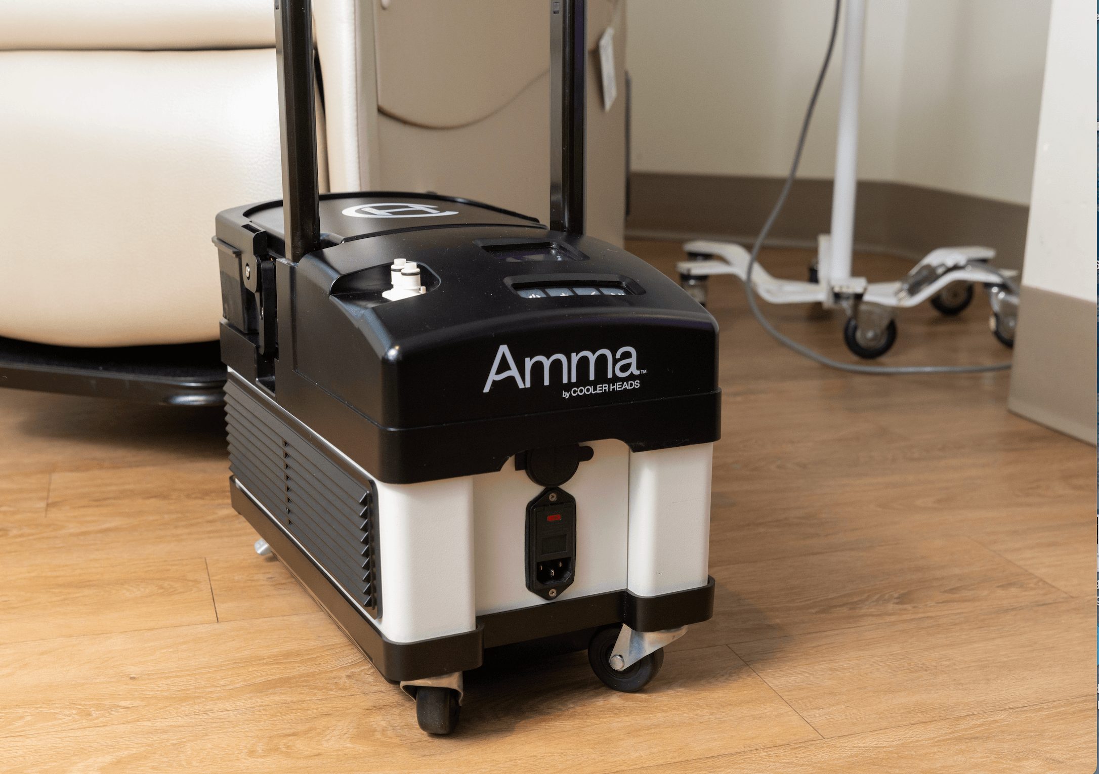 Medical device on wheels with black and white body, labeled 'Amma', on wooden floor near furniture and equipment.