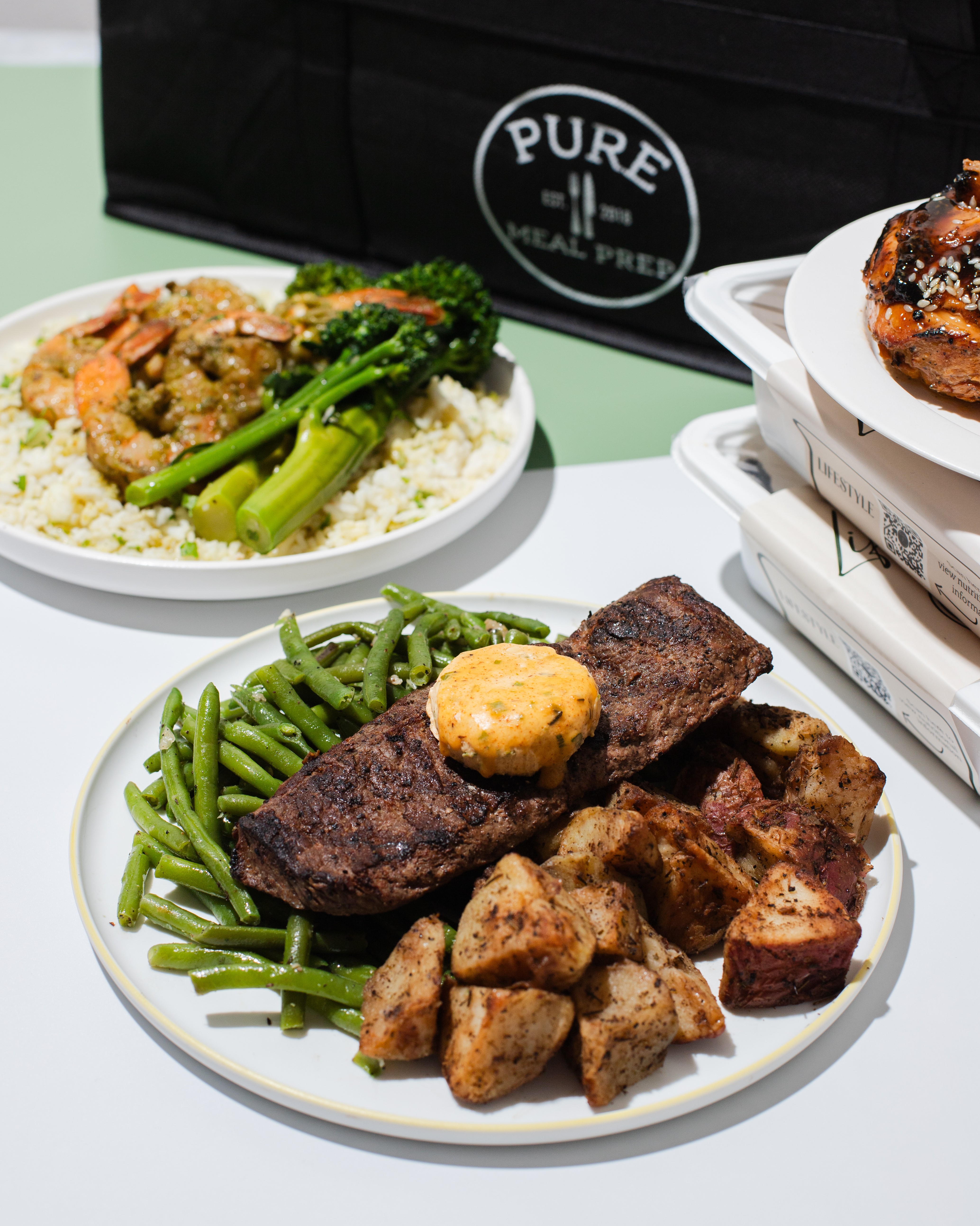 Plate with grilled steak, green beans, roasted potatoes, and a biscuit, on a white table with a black bag in background.