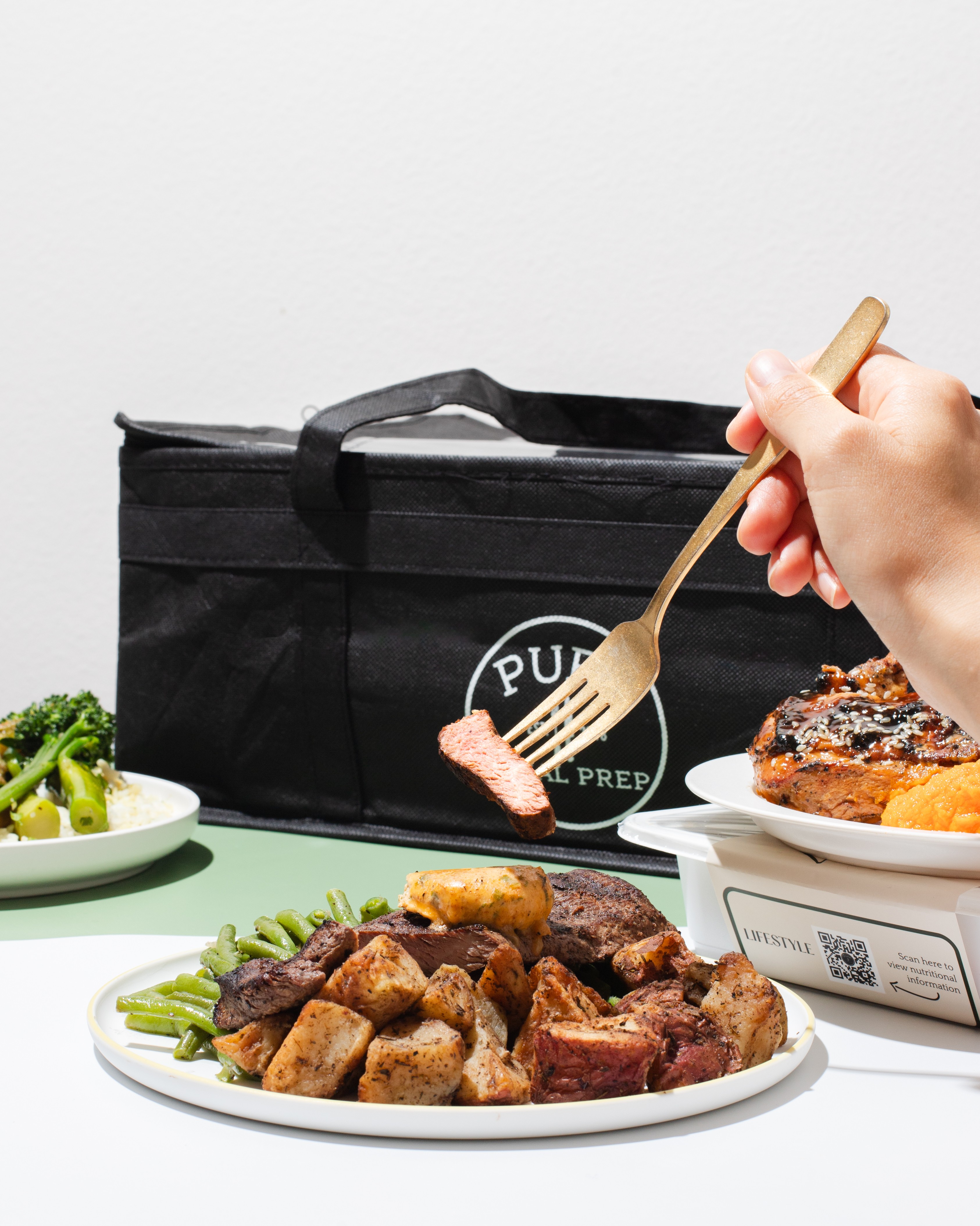 Hand holding a fork with food, black delivery bag, plate of cooked meat, and side dishes on a table.