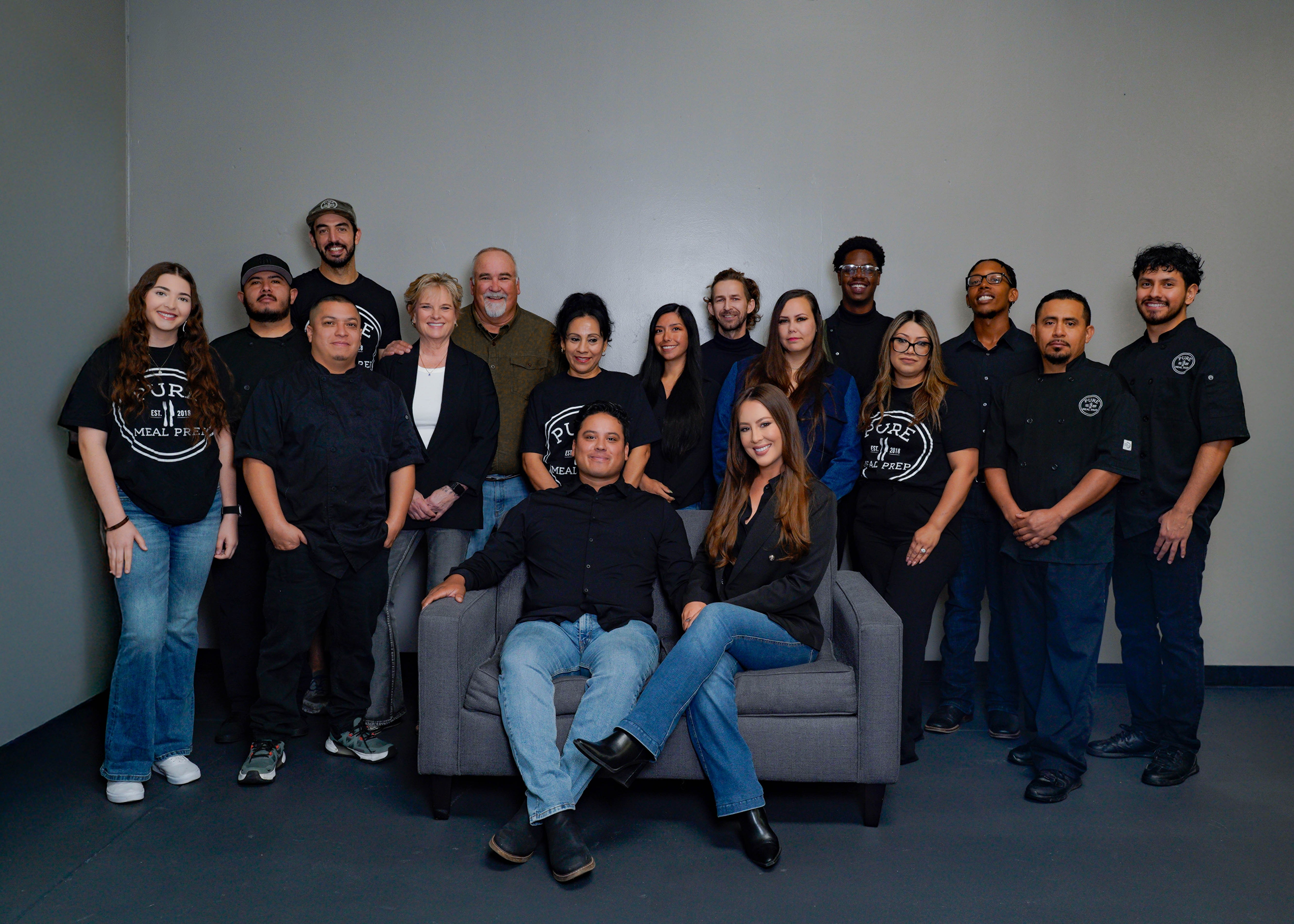 Group of 16 people, some standing and some sitting, in a room with a plain gray background.