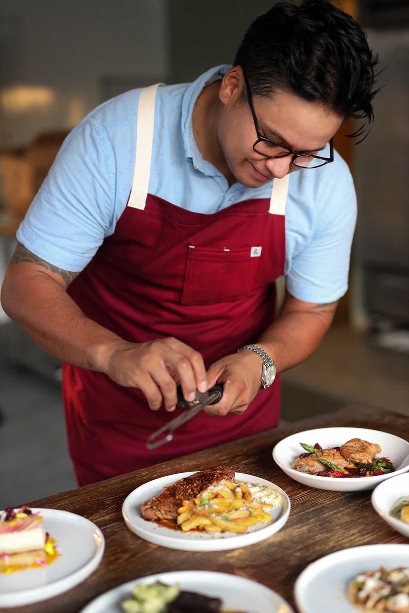 Man wearing glasses and a red apron preparing food in a kitchen, with dishes on the table in front of him.