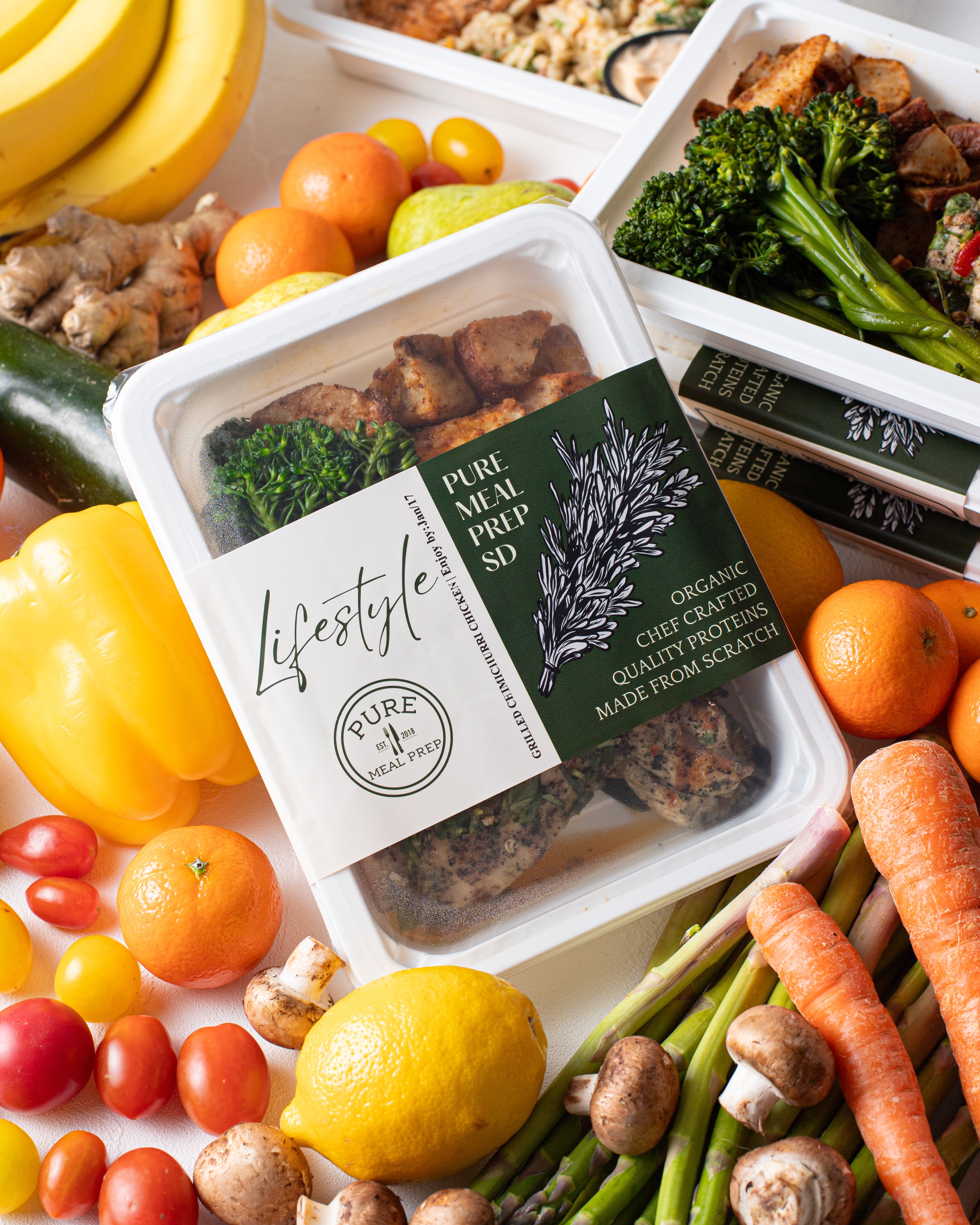 Assorted fresh fruits and vegetables surrounding packaged organic cooked chicken with broccoli, lemon, carrots, cherry tomatoes, bananas, and peppers.