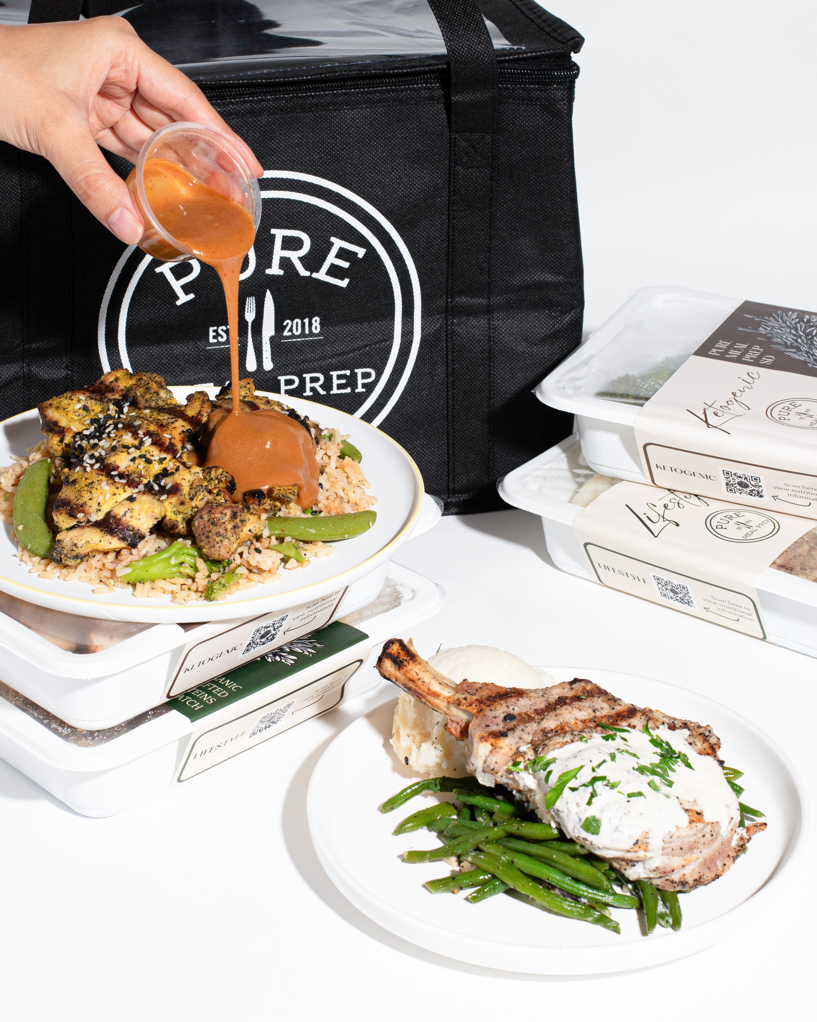 Hand pouring sauce over a burger on a plate, with takeout boxes and a black bag in the background.
