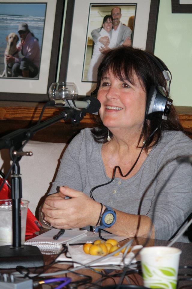 Woman with headphones smiling, sitting at a table with microphones, framed photos on the wall behind her.