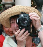 Person wearing a large straw hat taking a selfie with a camera, smiling, with another person partially visible behind.