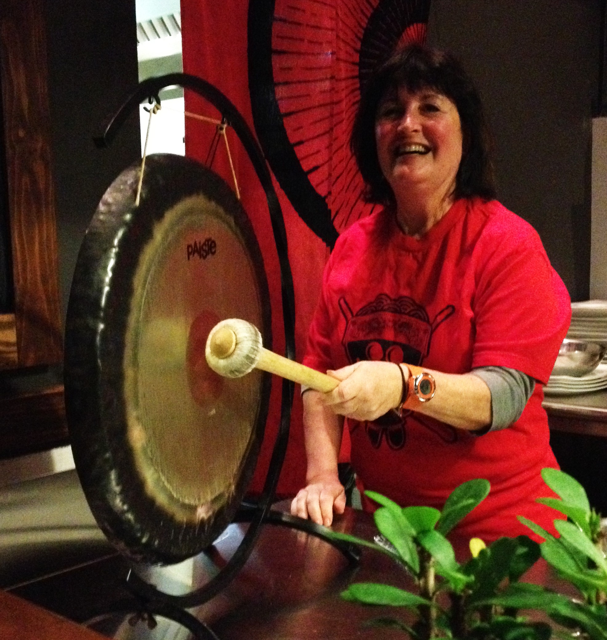 Woman smiling and playing a gong with a mallet, wearing a red shirt, in a room with red and black decor.