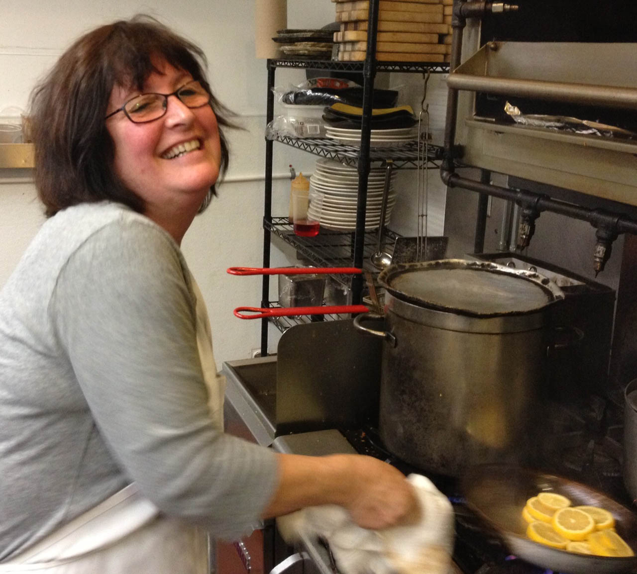 Woman smiling in a kitchen with pots, pans, and a rack of dishes behind her.