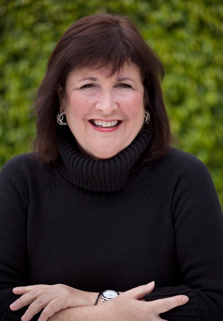 Smiling woman with dark hair wearing a black turtleneck sweater, outdoors with green foliage background.
