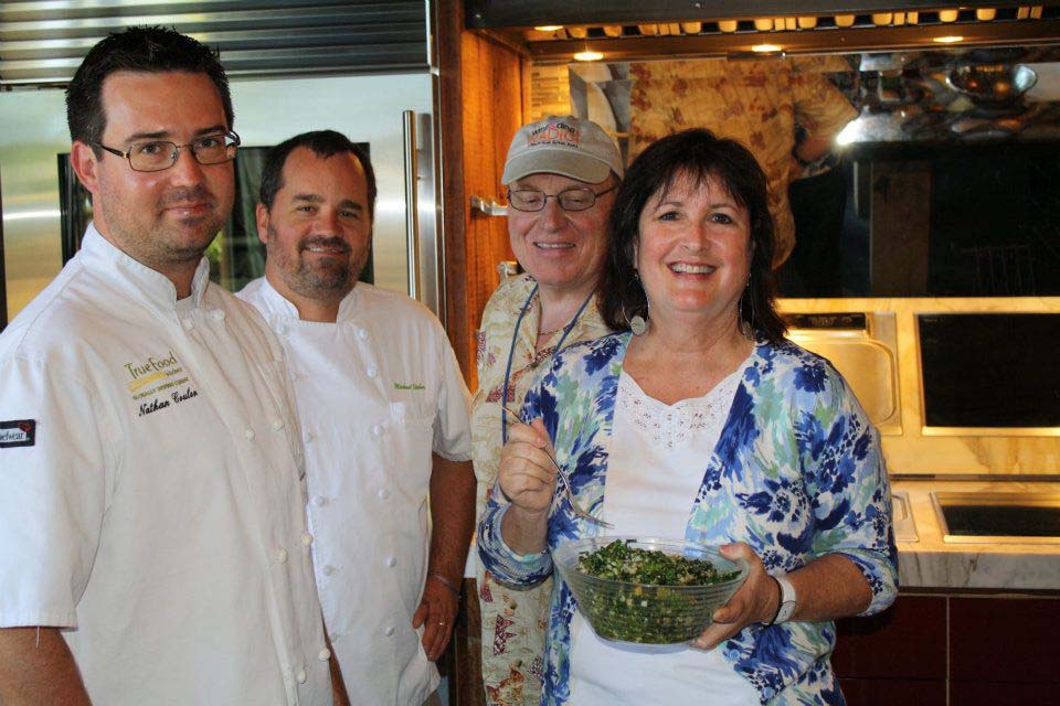 Four people smiling indoors, one holding a bowl of salad, with warm lighting and kitchen background.