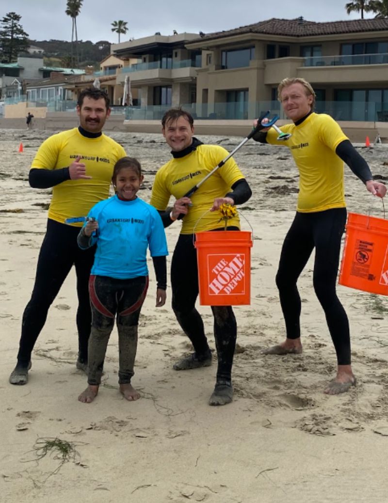 Four people, three adults and one child, standing on a beach with buildings in the background, holding buckets and tools, smiling.
