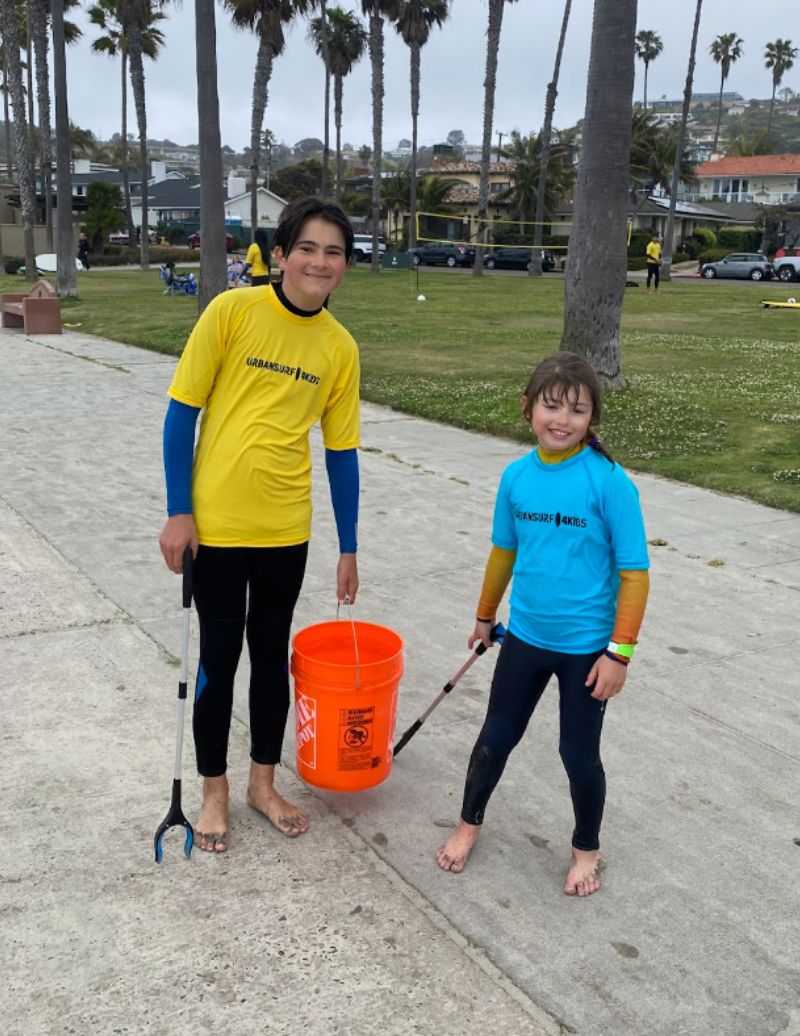 Two children standing on a sidewalk, one holding an orange bucket, with palm trees and a park in the background.