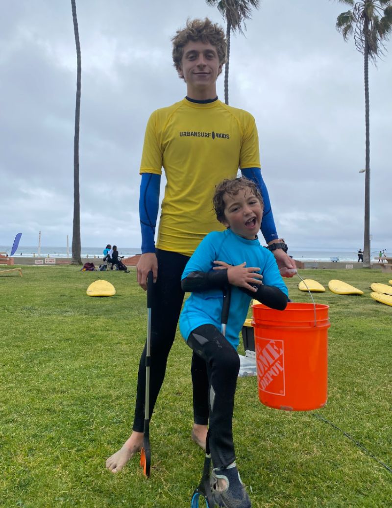 Two children outdoors on grass, one holding an orange bucket, with palm trees and cloudy sky in background.