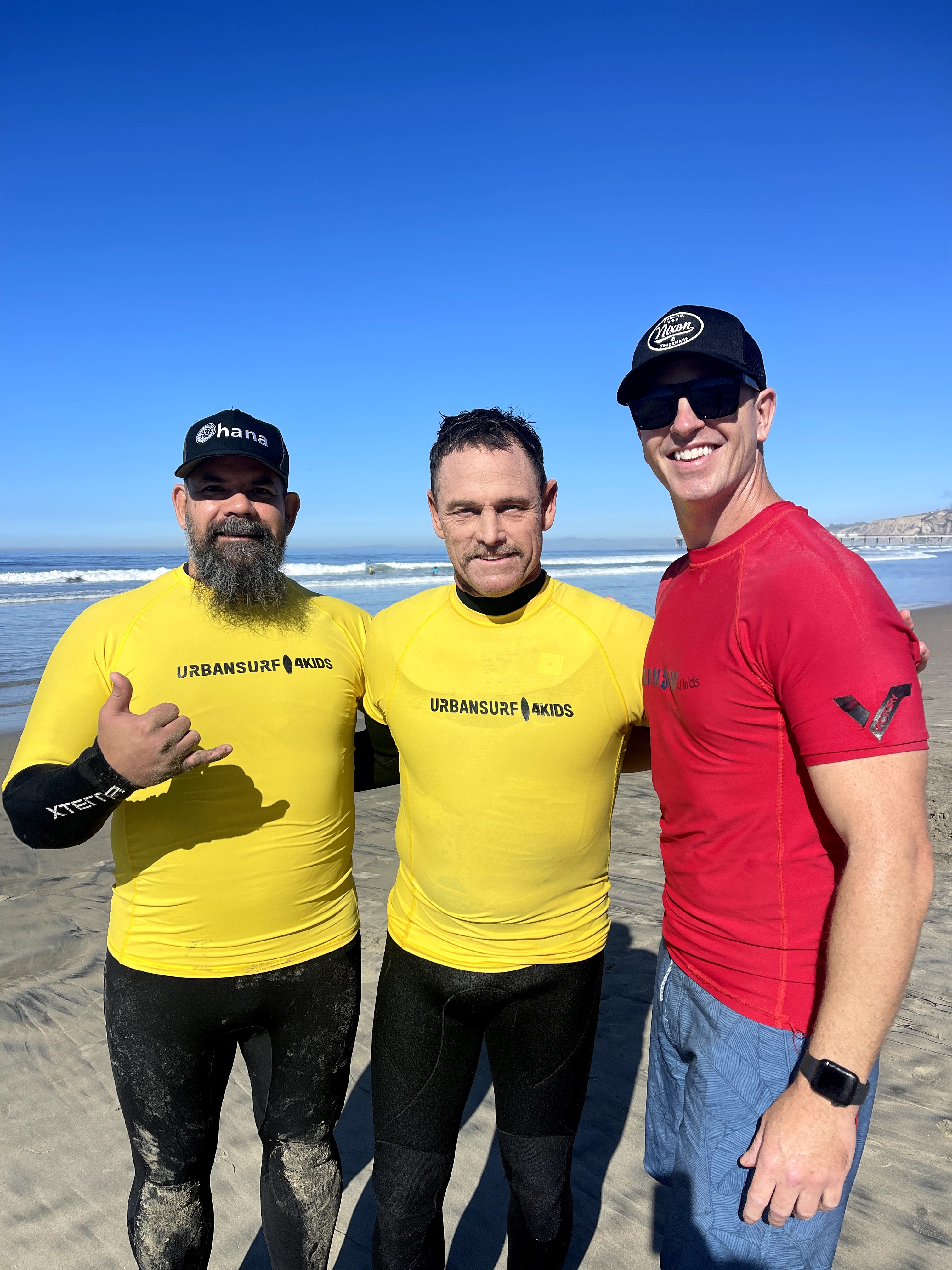 Three men standing on a beach with blue sky, wearing casual clothes, smiling, with ocean waves in background.