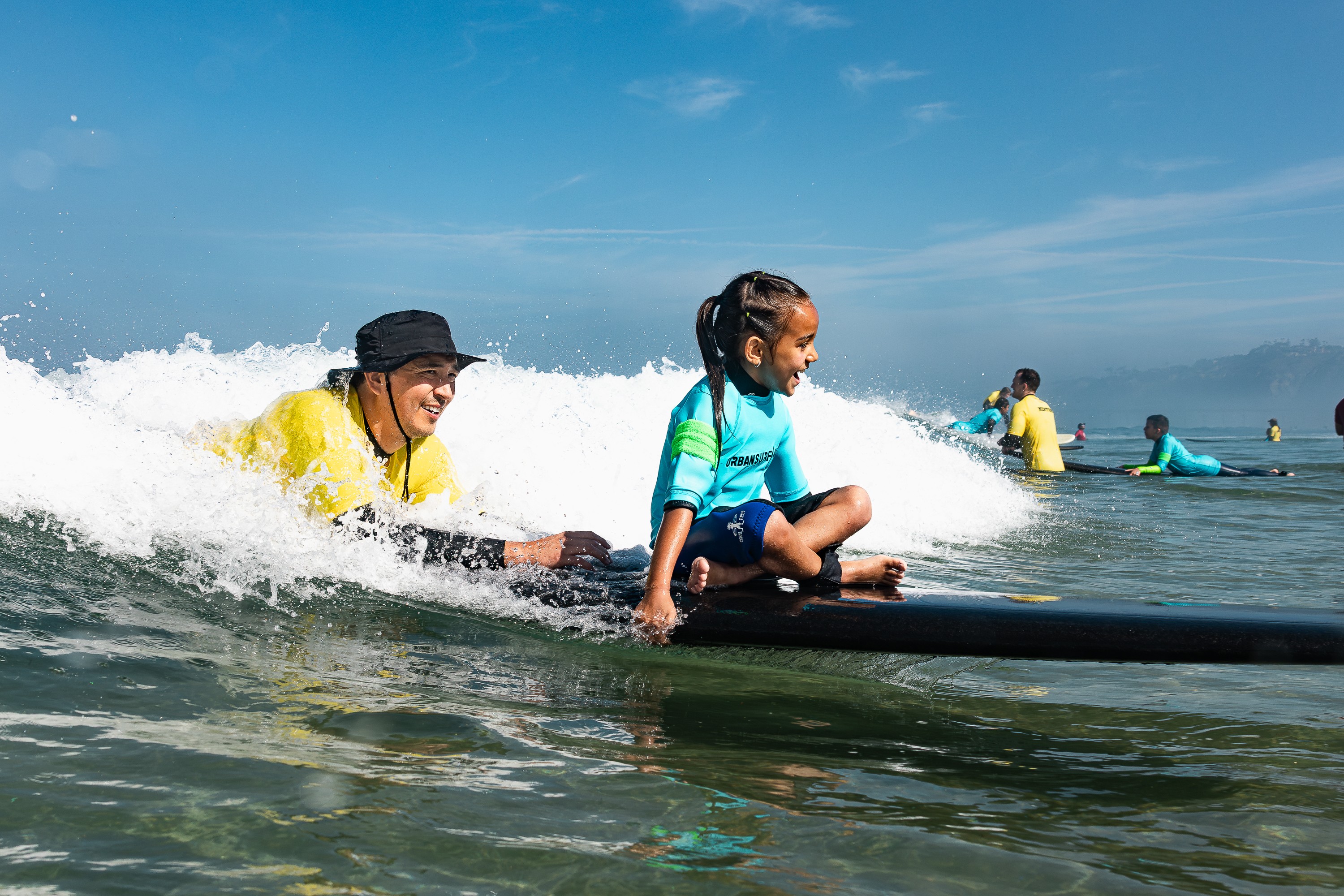 Children learning to surf in the ocean with waves, wearing life jackets and wetsuits, under a blue sky.