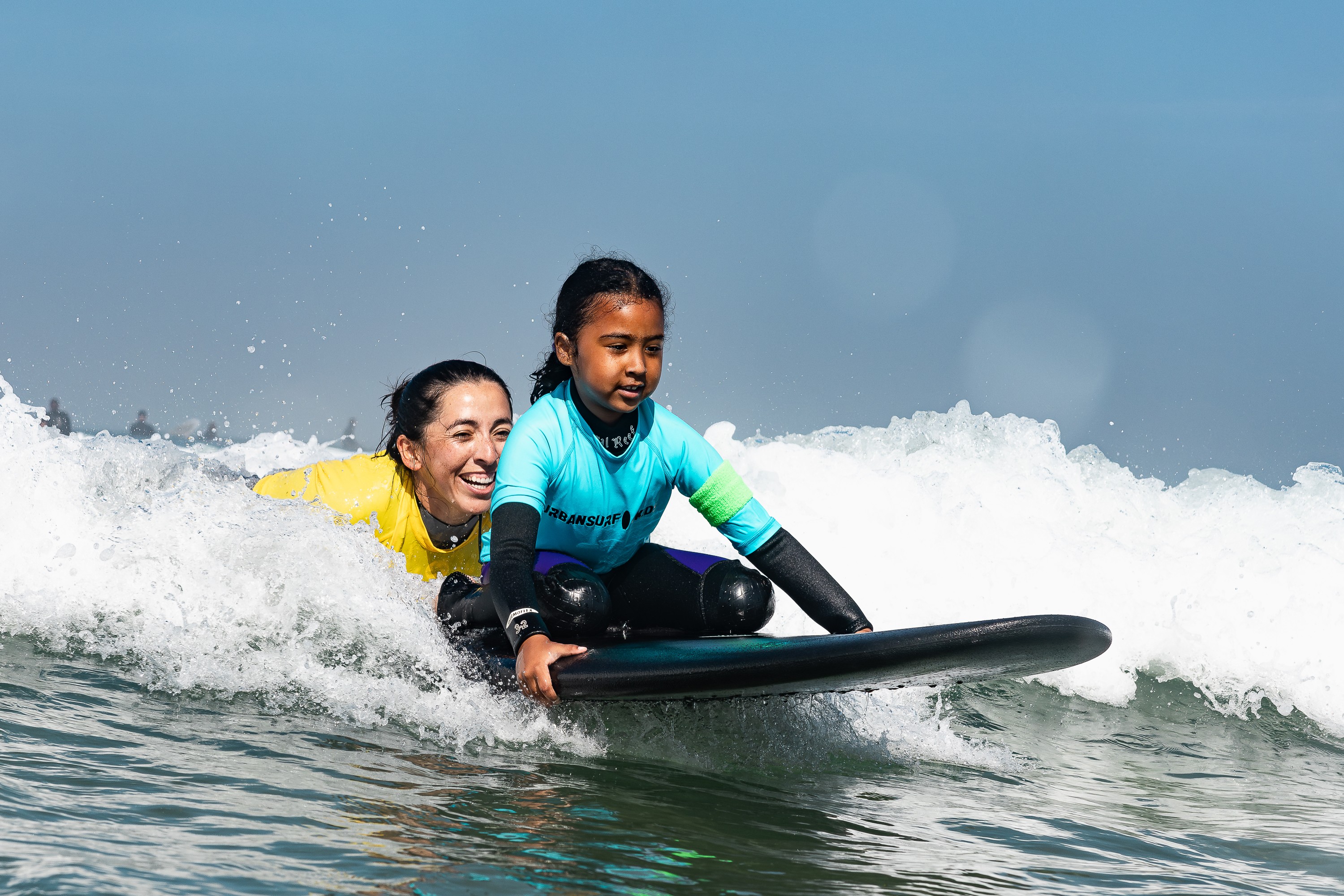 Two children riding a surfboard on a wave, smiling, with a clear blue sky background.