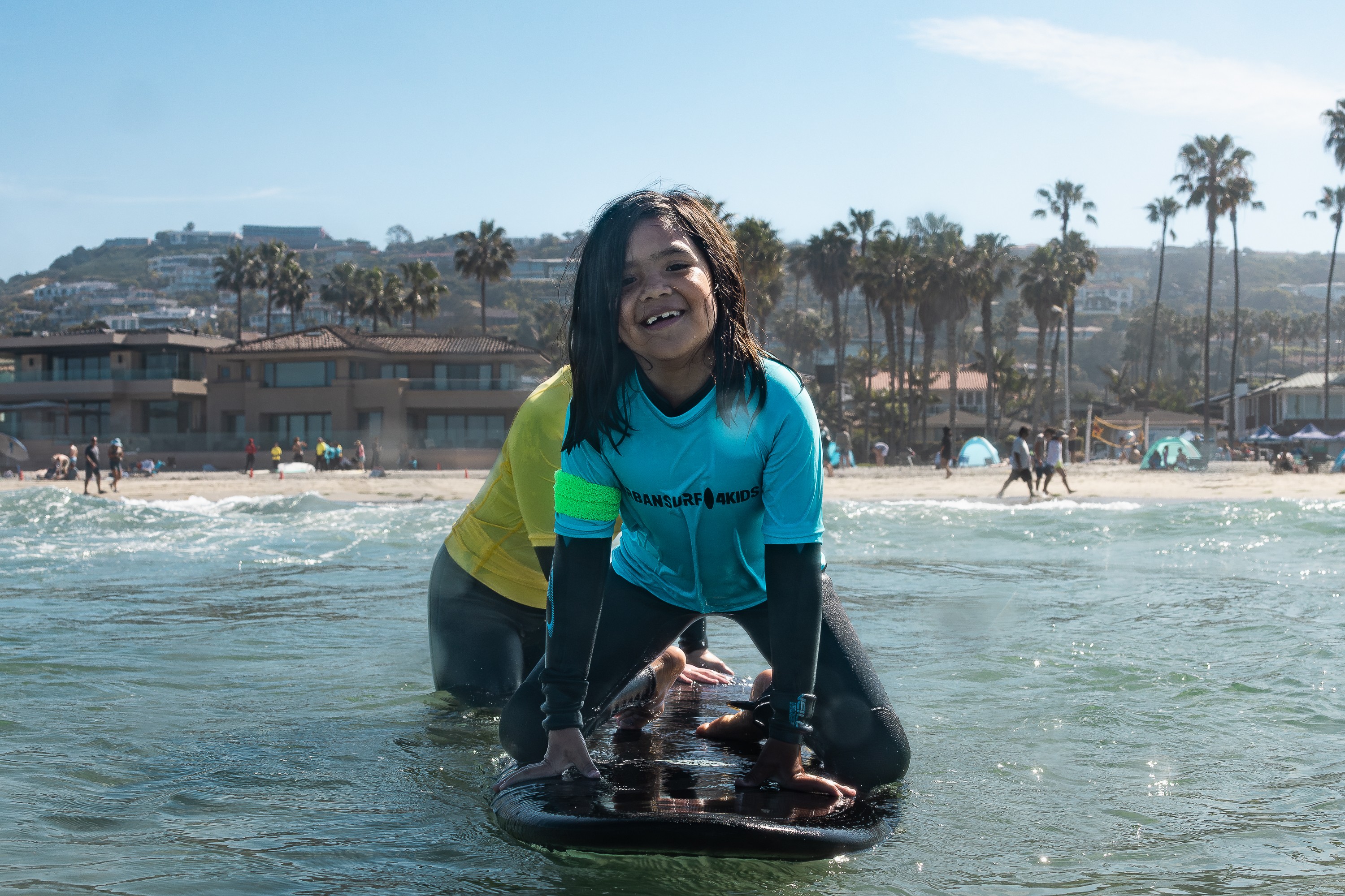 Person smiling on a surfboard in the water at the beach with palm trees and buildings in the background.