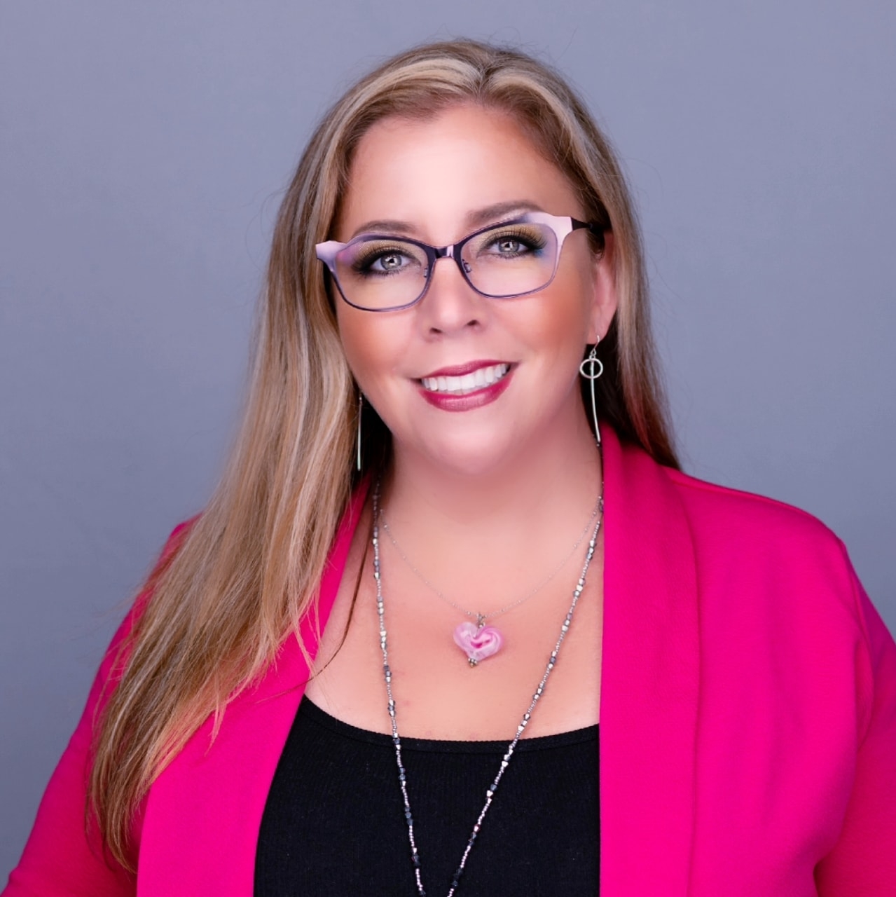 Woman with long blonde hair, glasses, pink blazer, black top, smiling, against gray background.