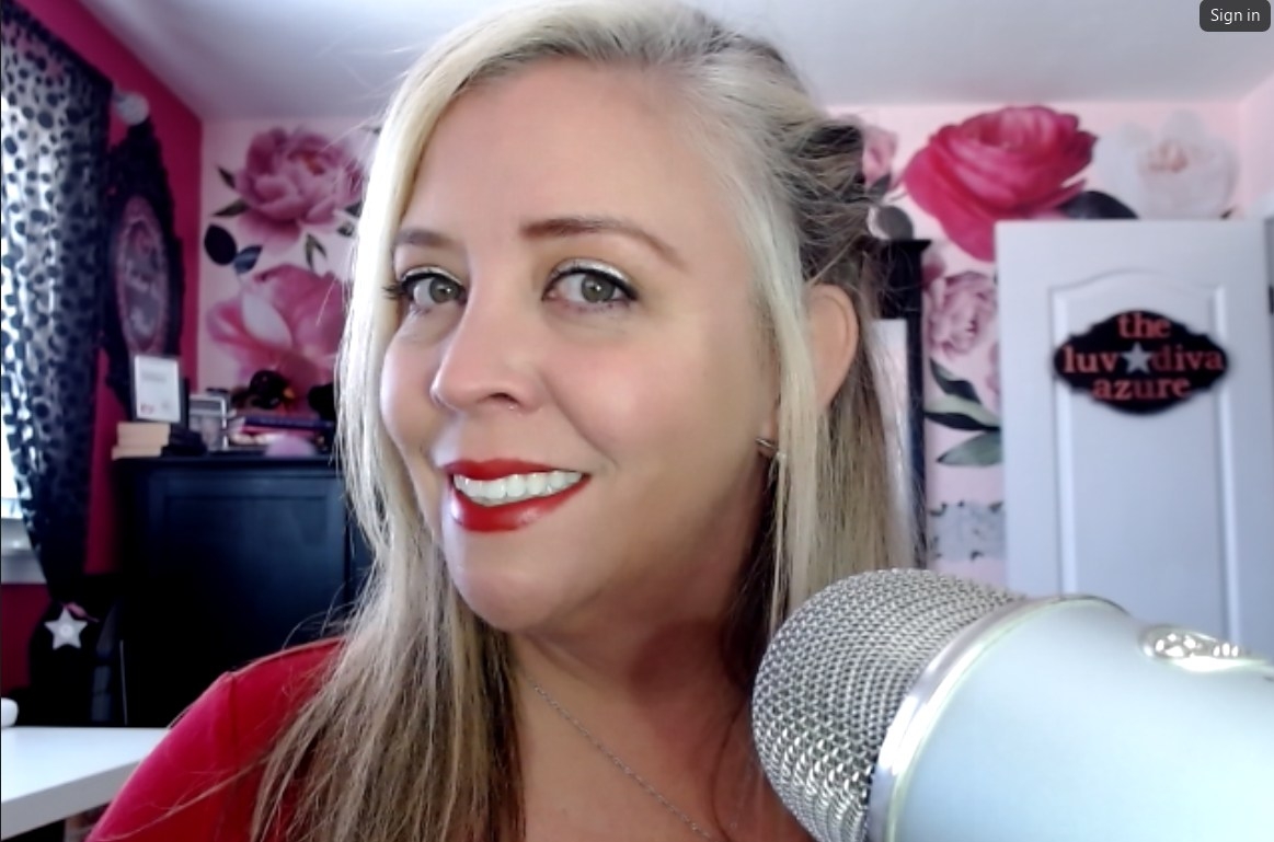 Close-up of a smiling woman with long blonde hair in a room with floral wallpaper, a microphone in foreground.