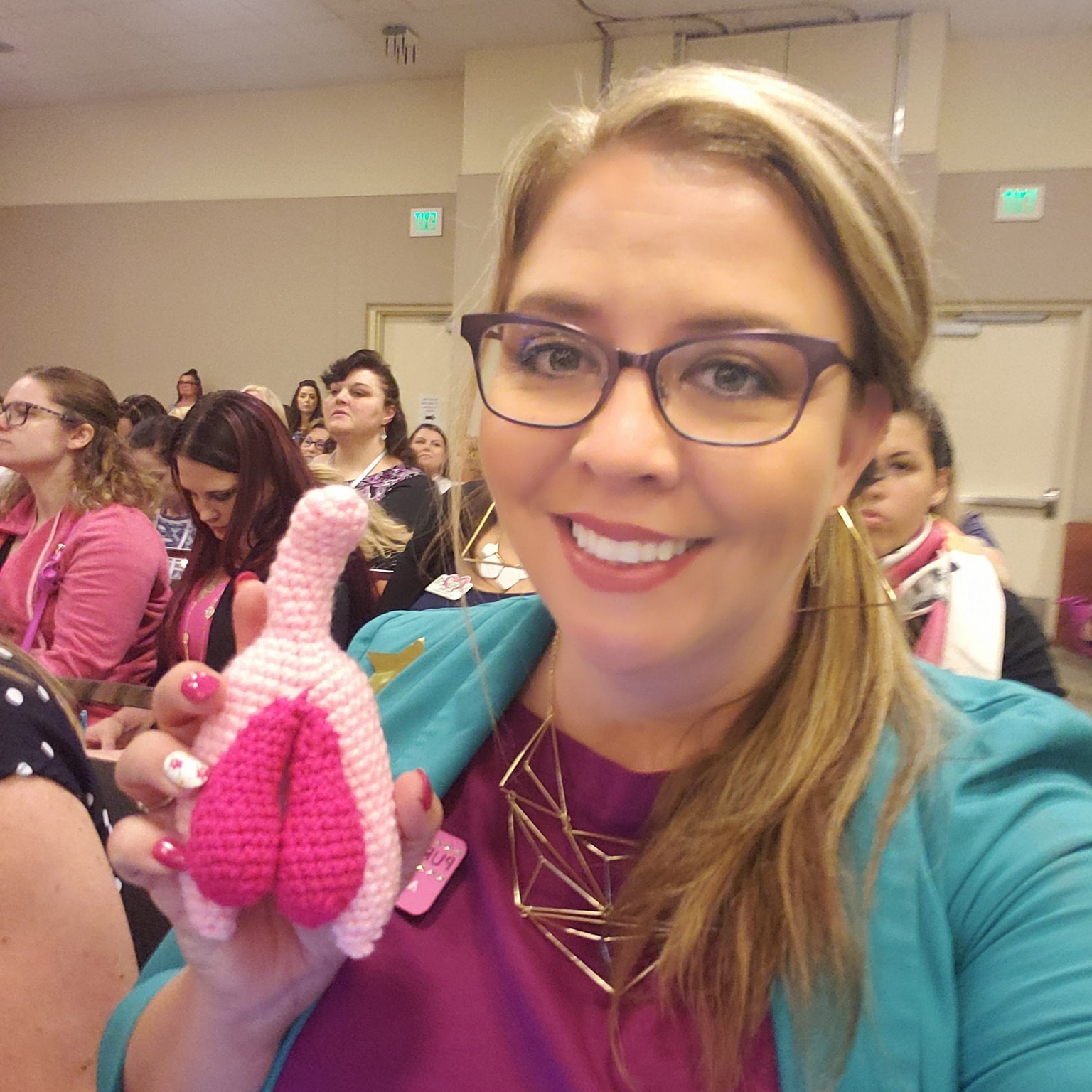 Woman with glasses smiling, holding pink and white knitted object, in a room with seated people in the background.