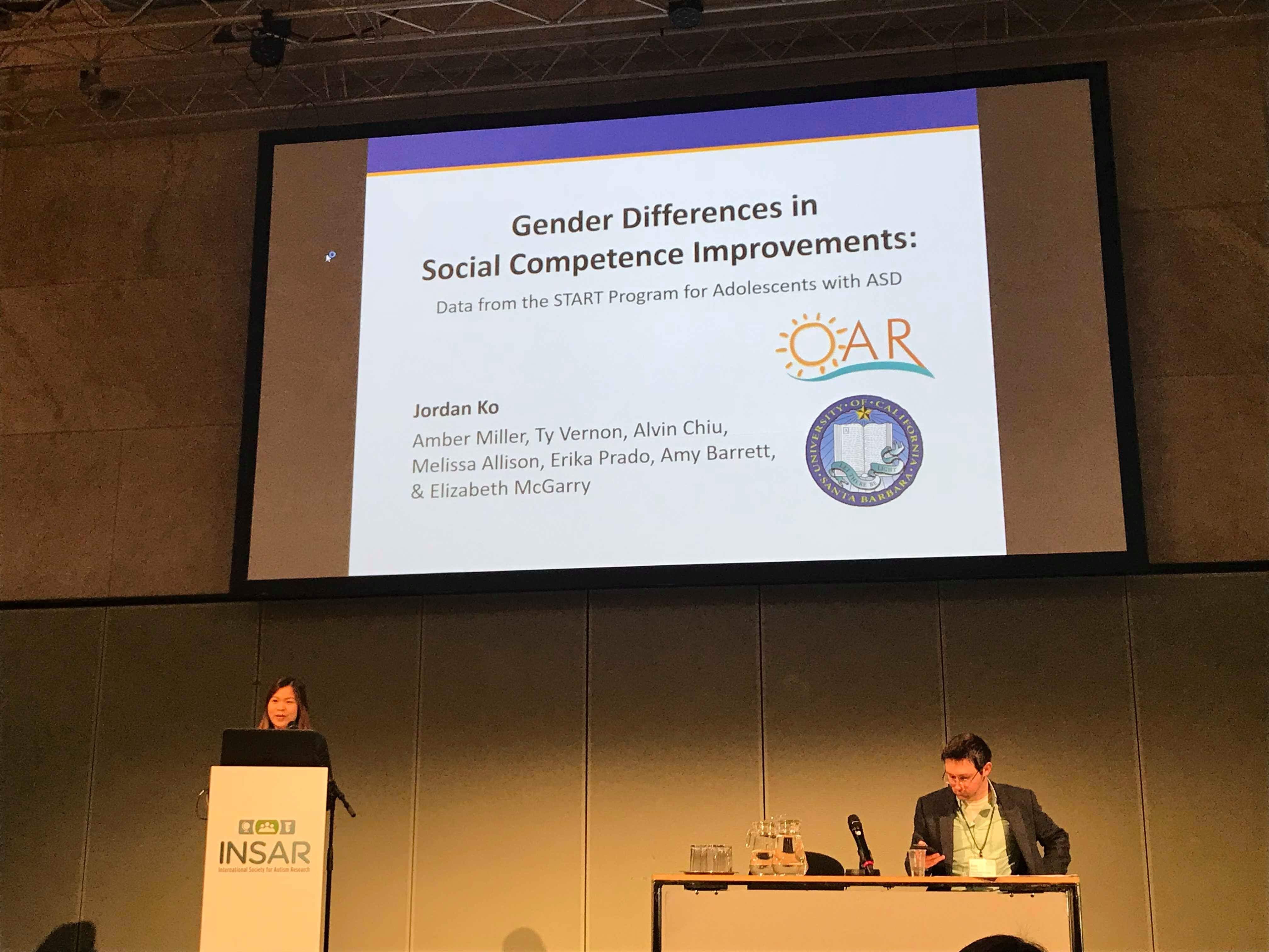 Presentation slide titled 'Gender Differences in Social Competence Improvements' with four authors' names and logos, in a conference room.