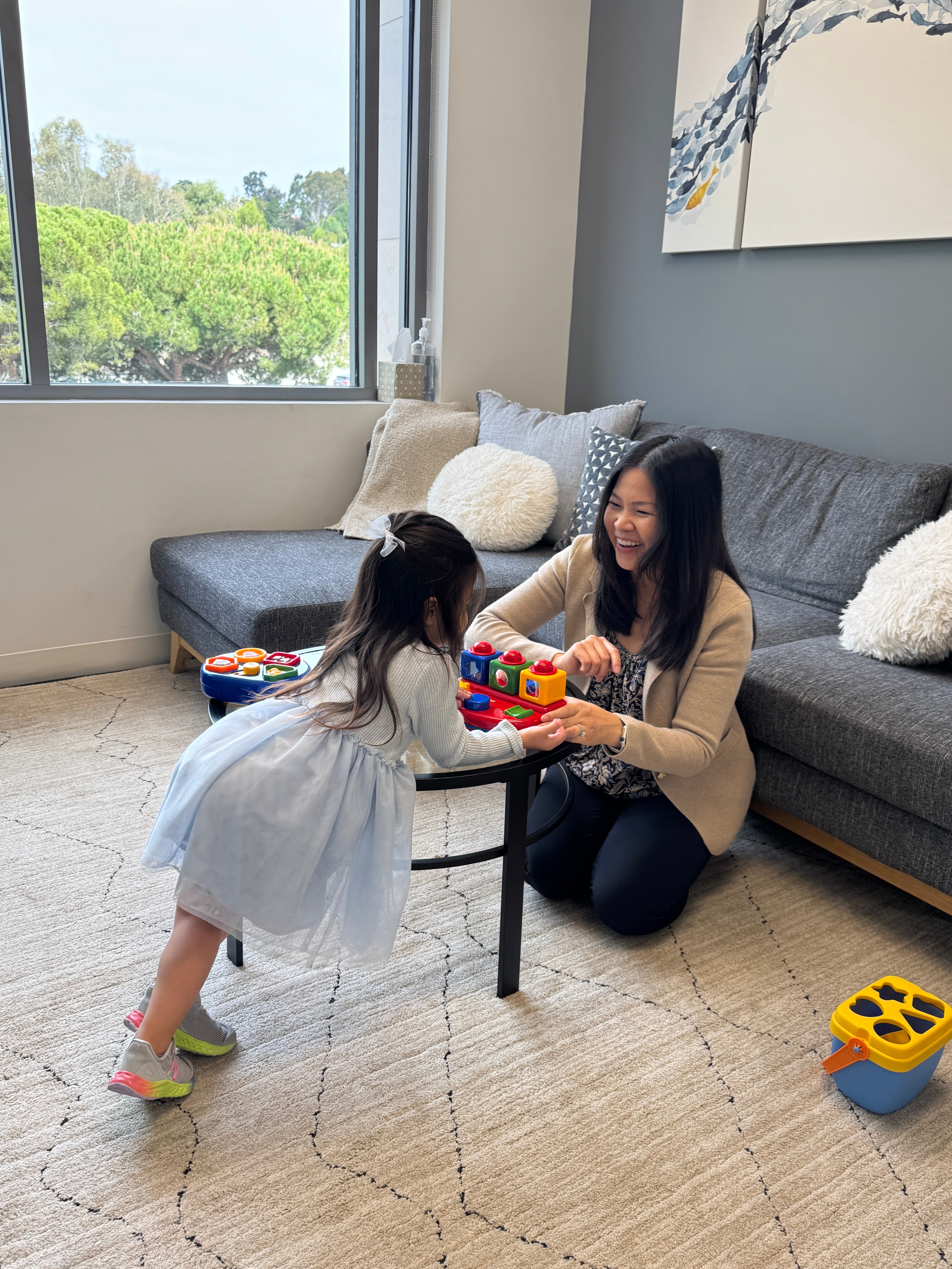 A woman and a young girl play with colorful toys on a small table in a living room, near a window with a view of trees.