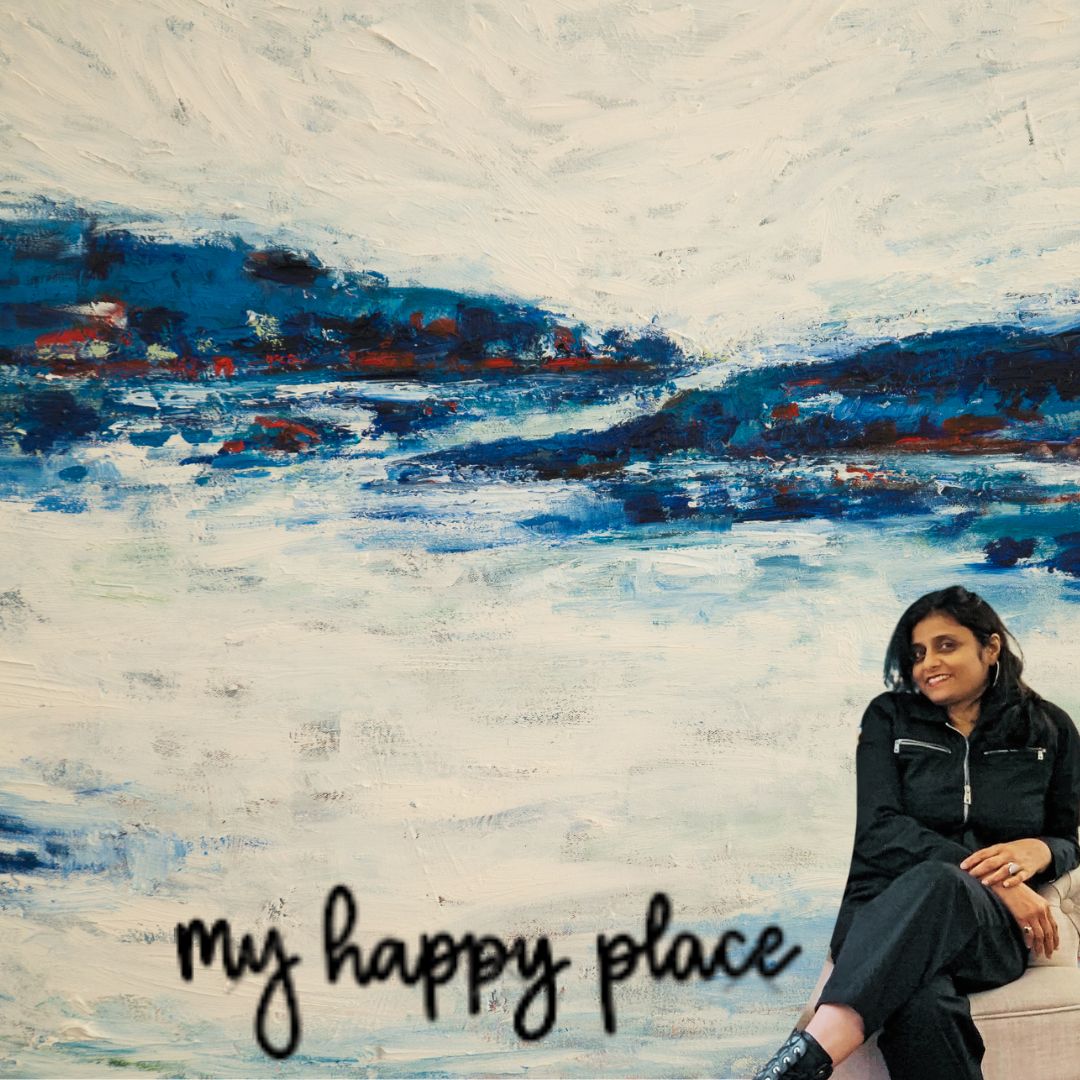 A woman sitting outdoors near a large abstract landscape painting with the text 'my happy place' below.