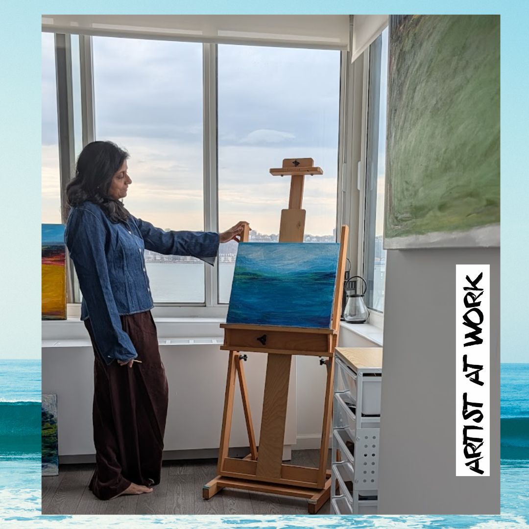 Woman painting a seascape on an easel indoors near a large window, with a green painting on the wall. Word 'ARTIST AT WORK' on side.