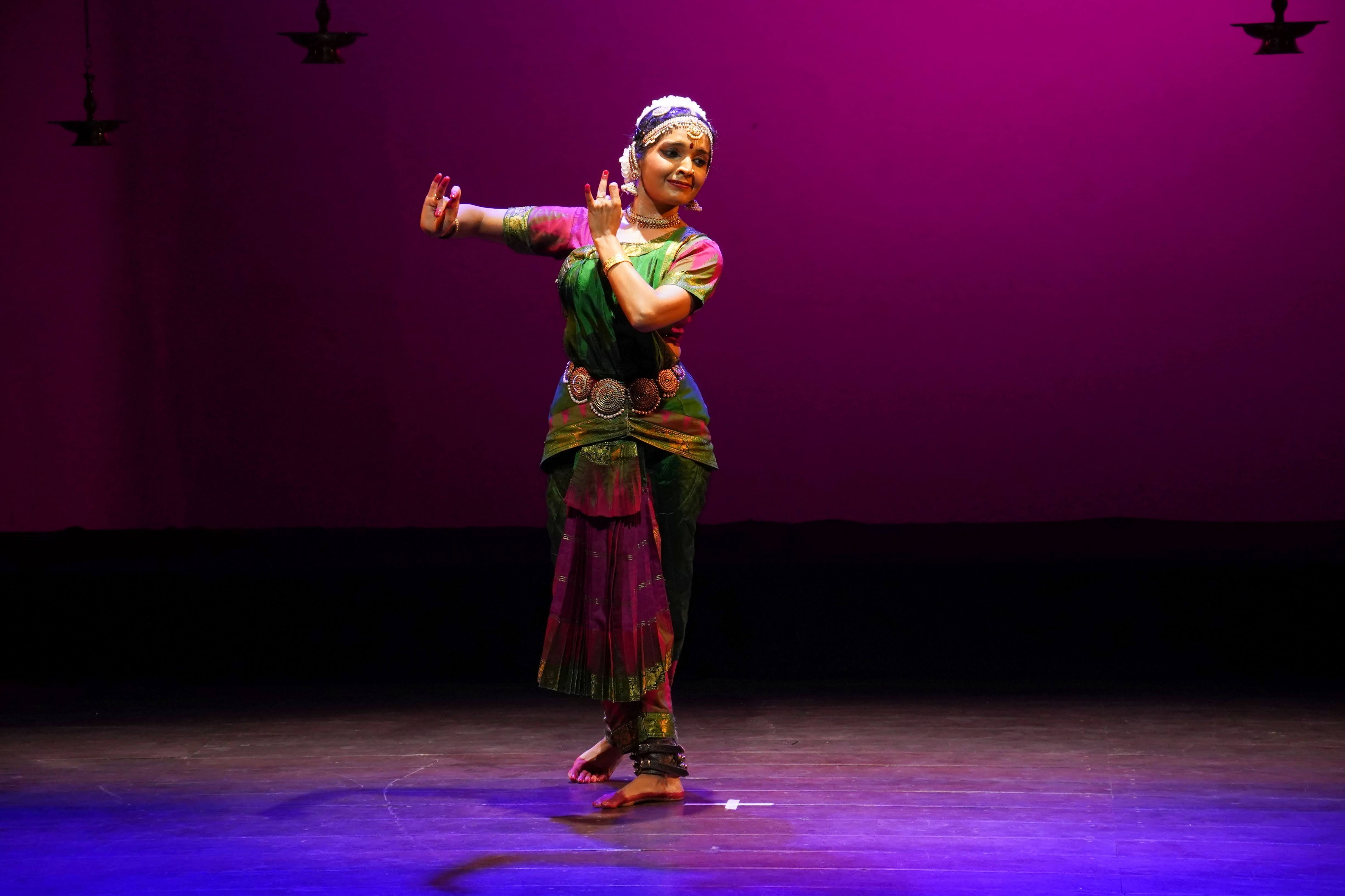 Performer in traditional costume dancing on stage with purple background, arms extended, focused expression.