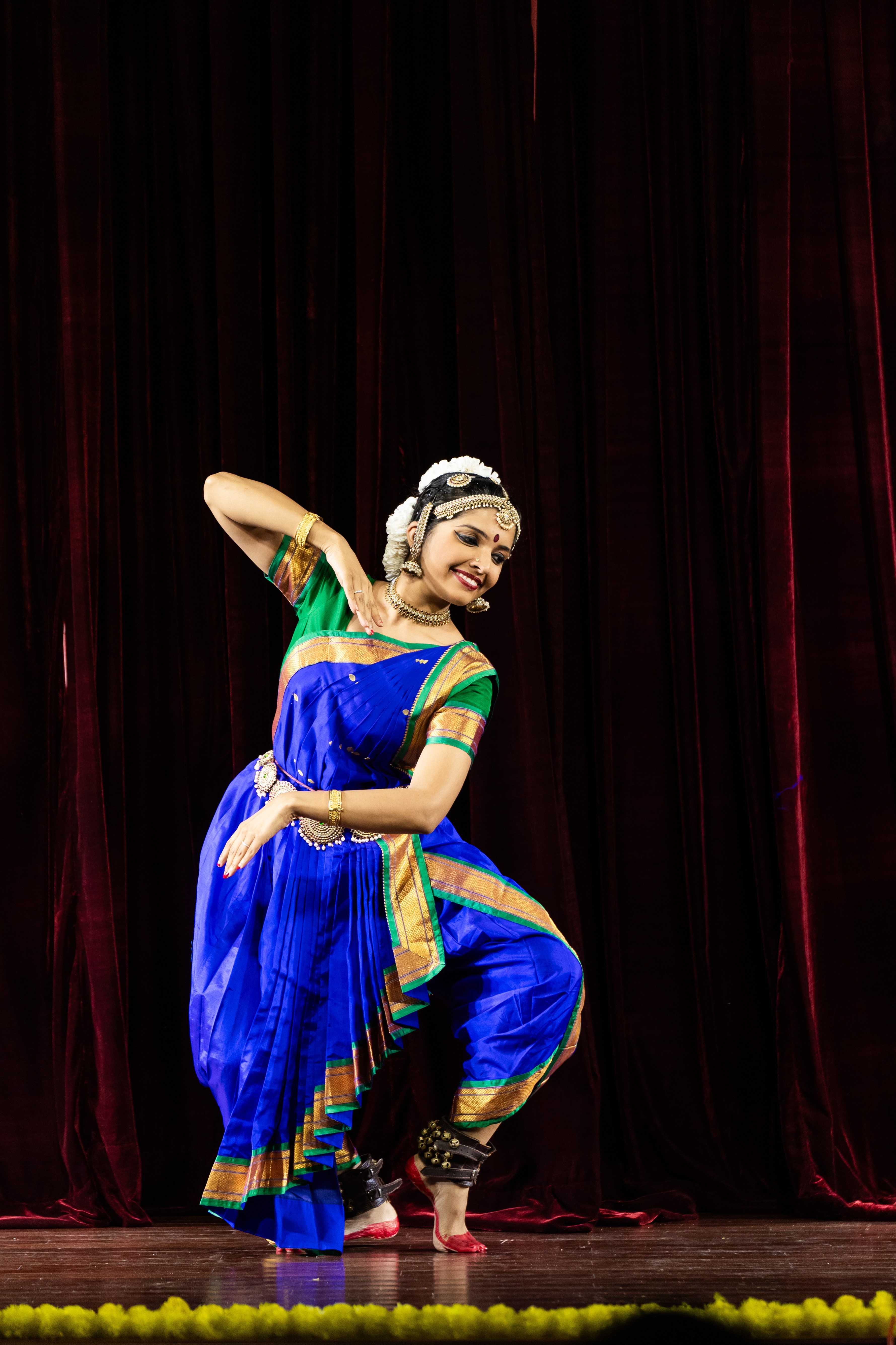 A woman in a blue and green traditional dance costume performs on stage with a dark curtain background.