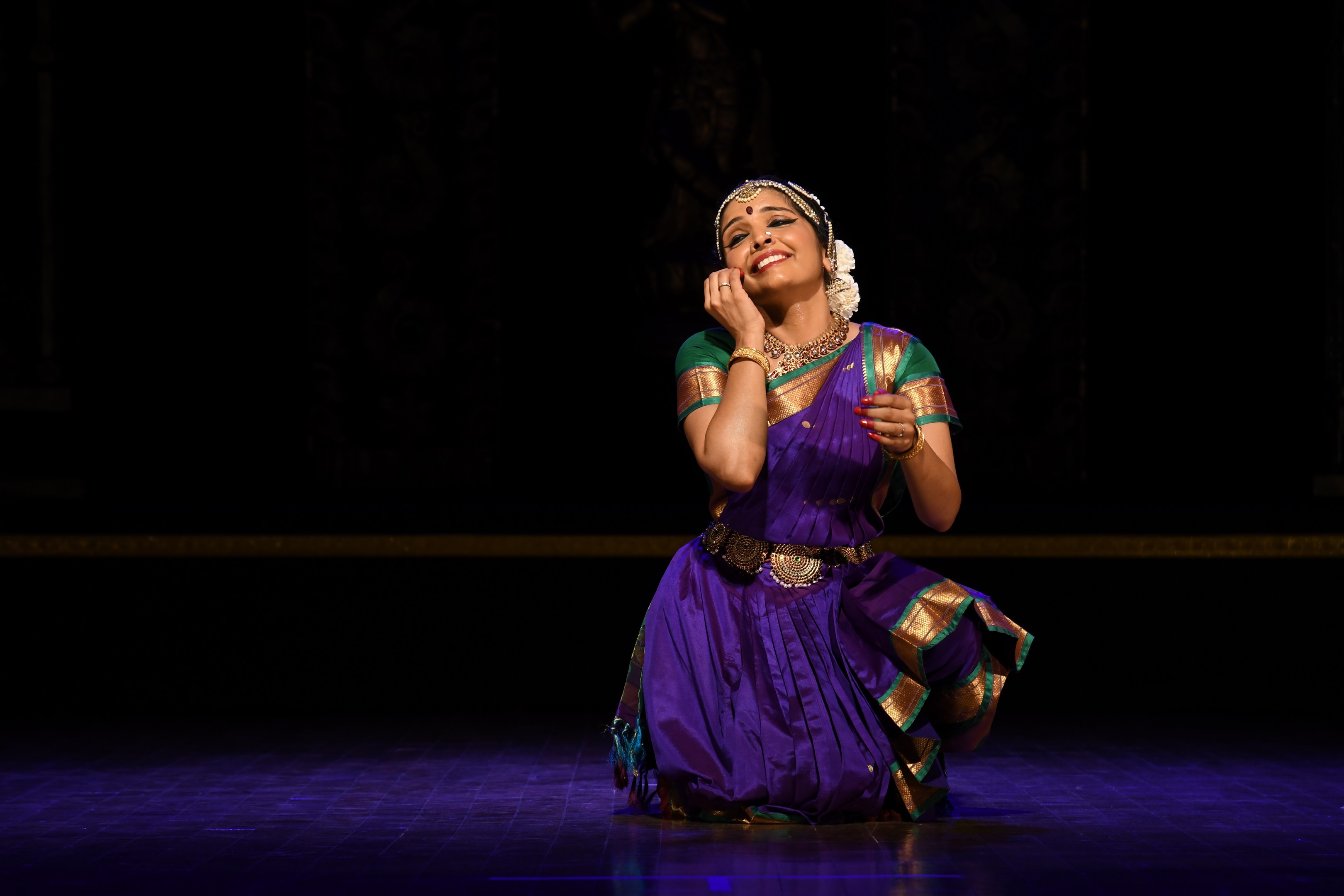 A woman in traditional attire kneels on stage, smiling with hands near her face, against a dark background.
