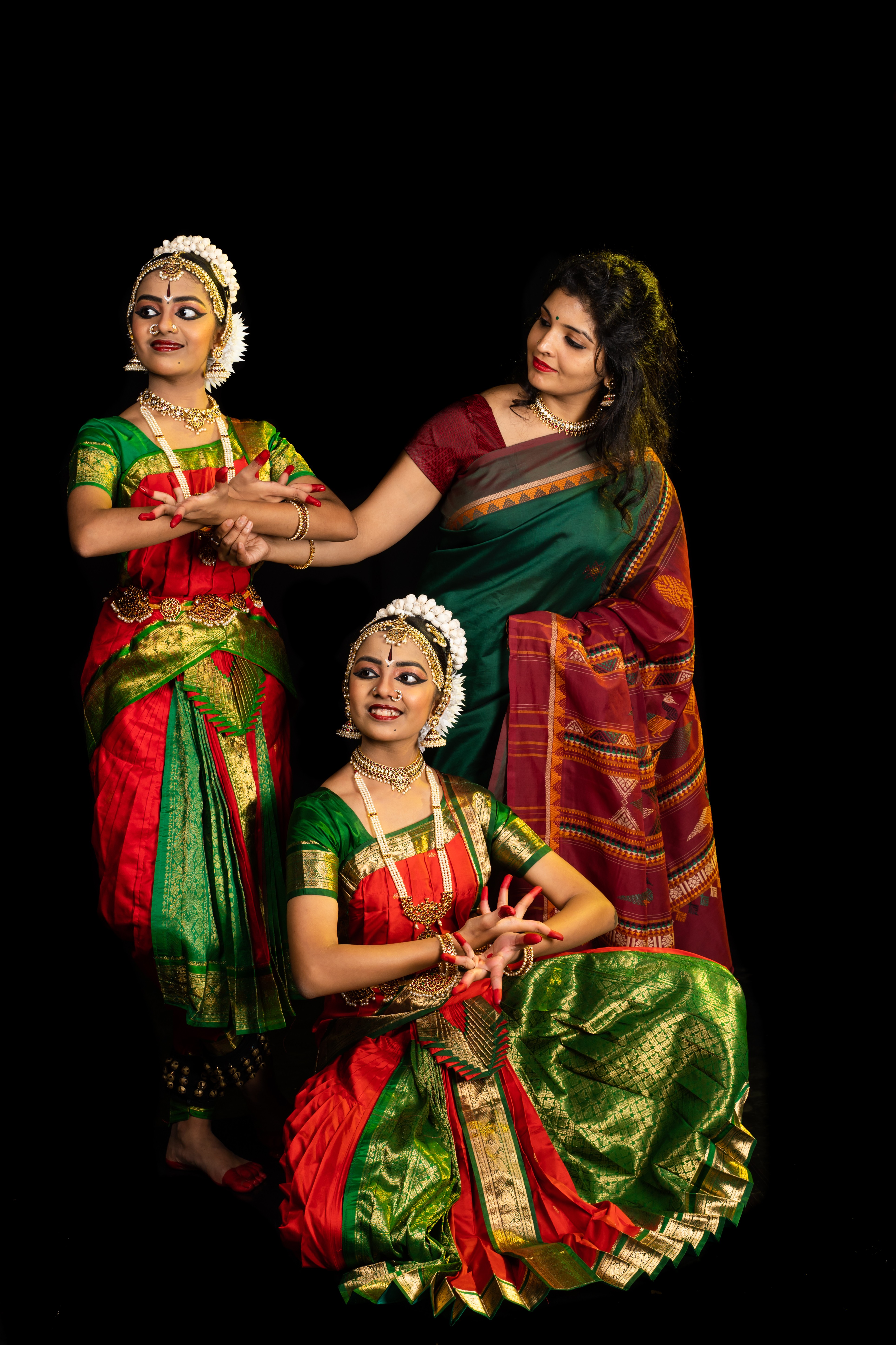 Three women in traditional colorful Indian attire, two standing and one sitting, against a black background.