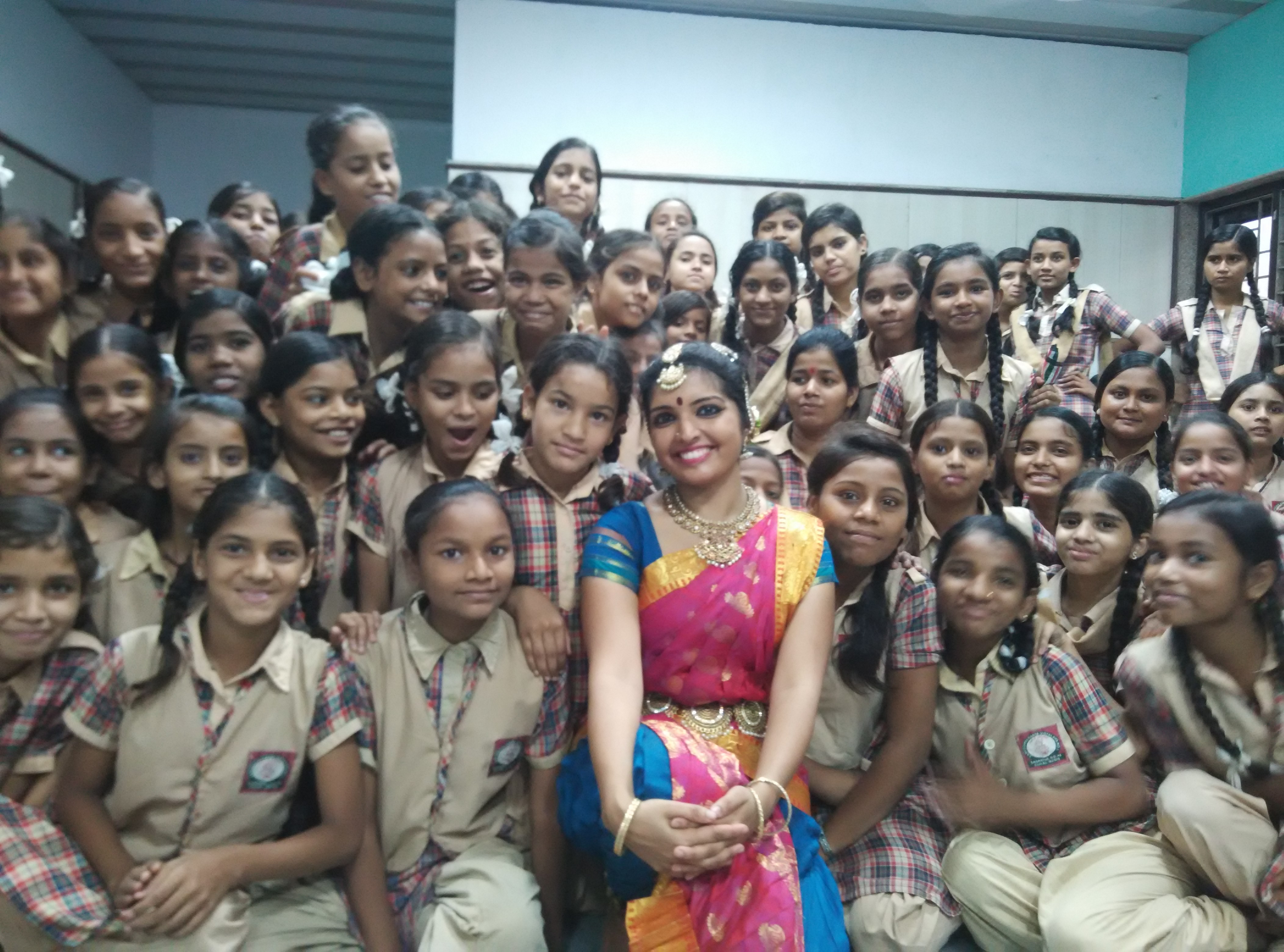 Group of school children with a woman in colorful traditional attire, smiling in classroom setting.