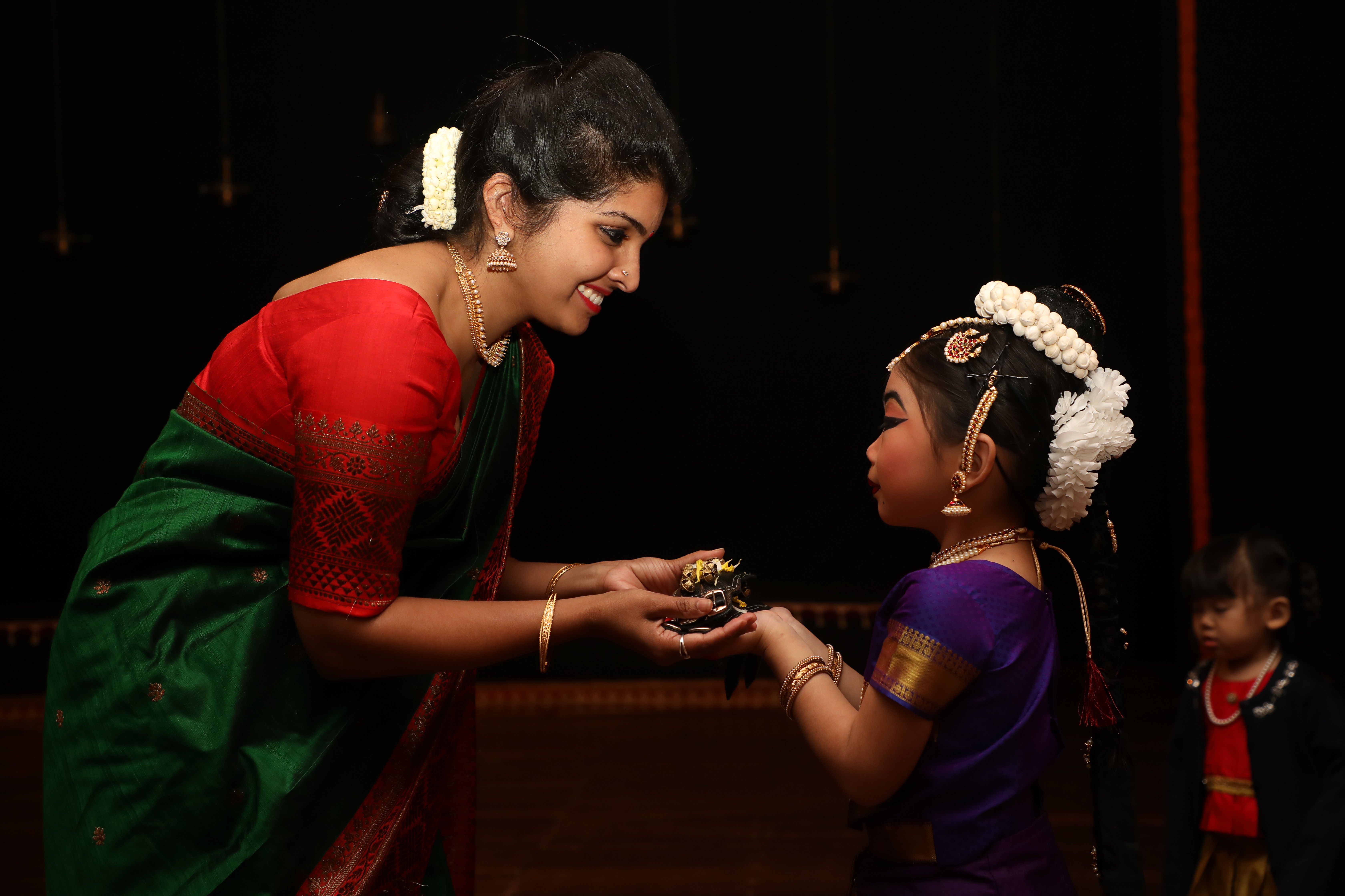 Woman in a green and red saree hands a gift to a girl in a purple dress with white hair accessories, on stage.