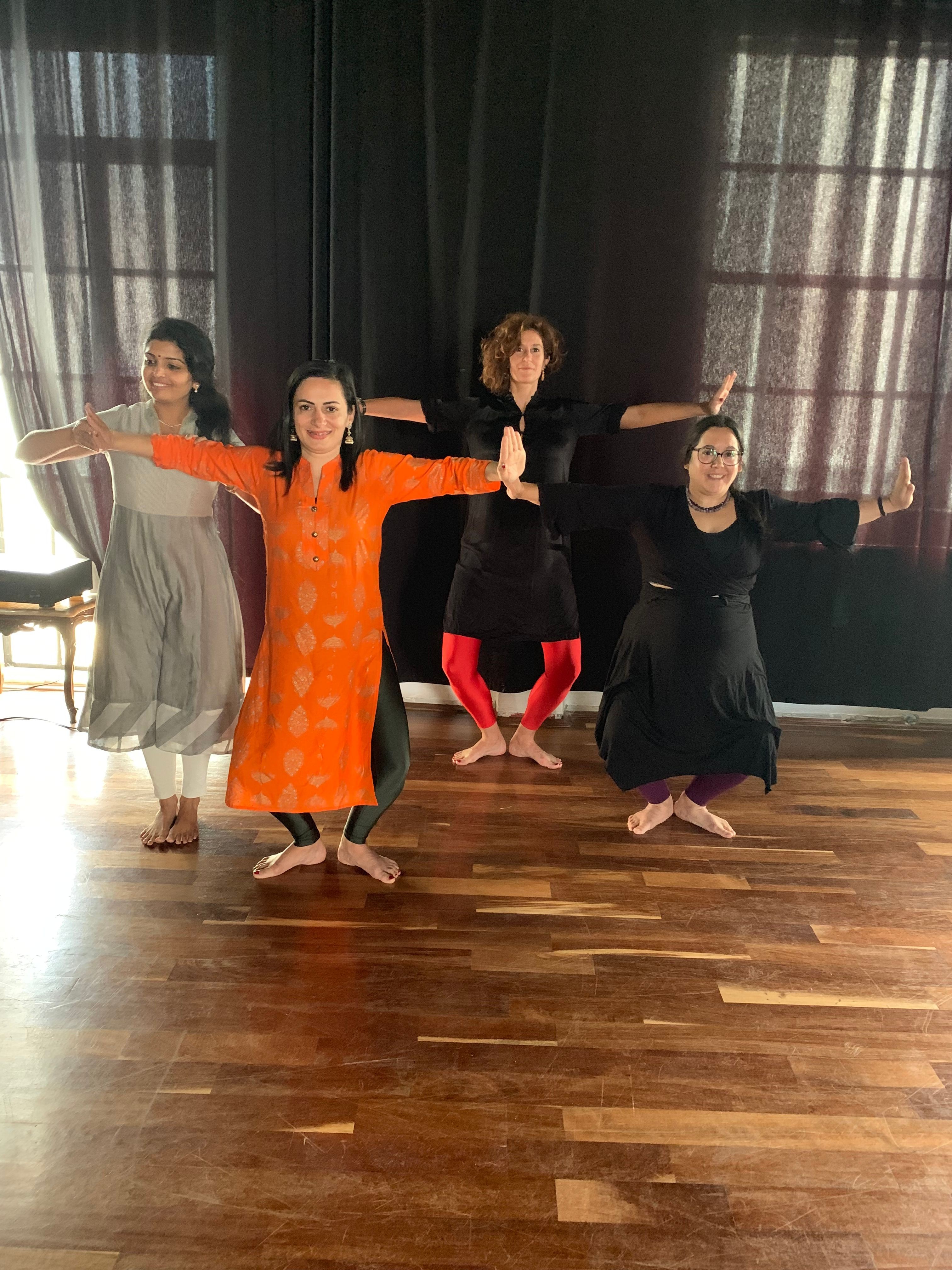 Four women standing on a wooden floor in front of a black curtain, one woman in an orange dress extending arms, others in black outfits.