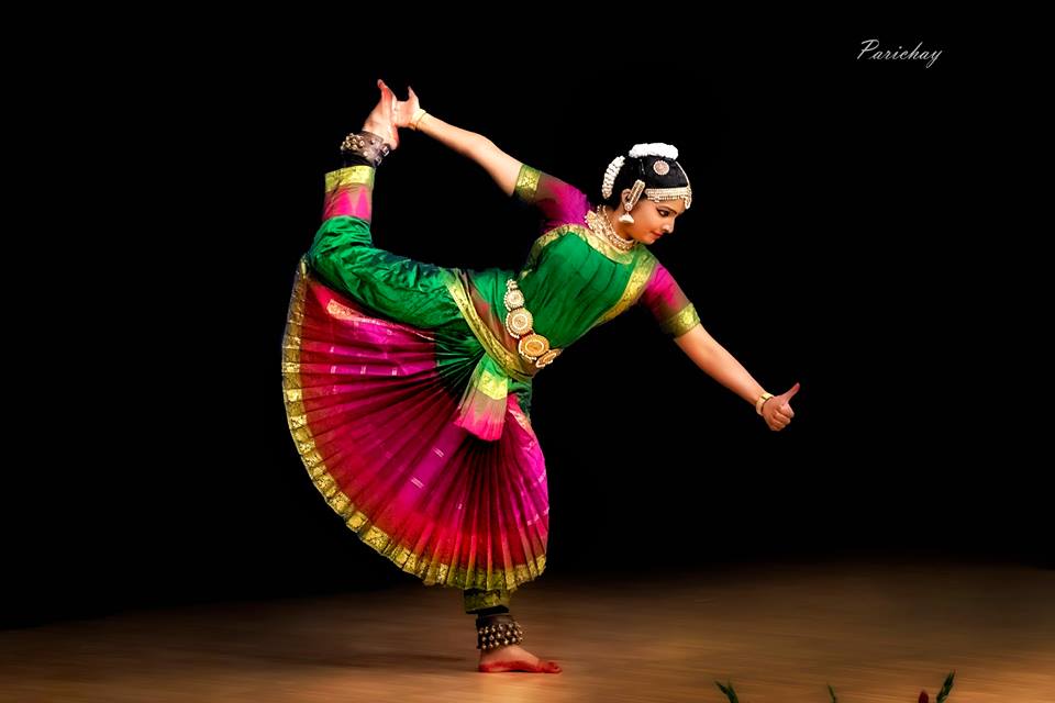 A classical Indian dancer in a green and pink costume performs a pose on stage with a black background.