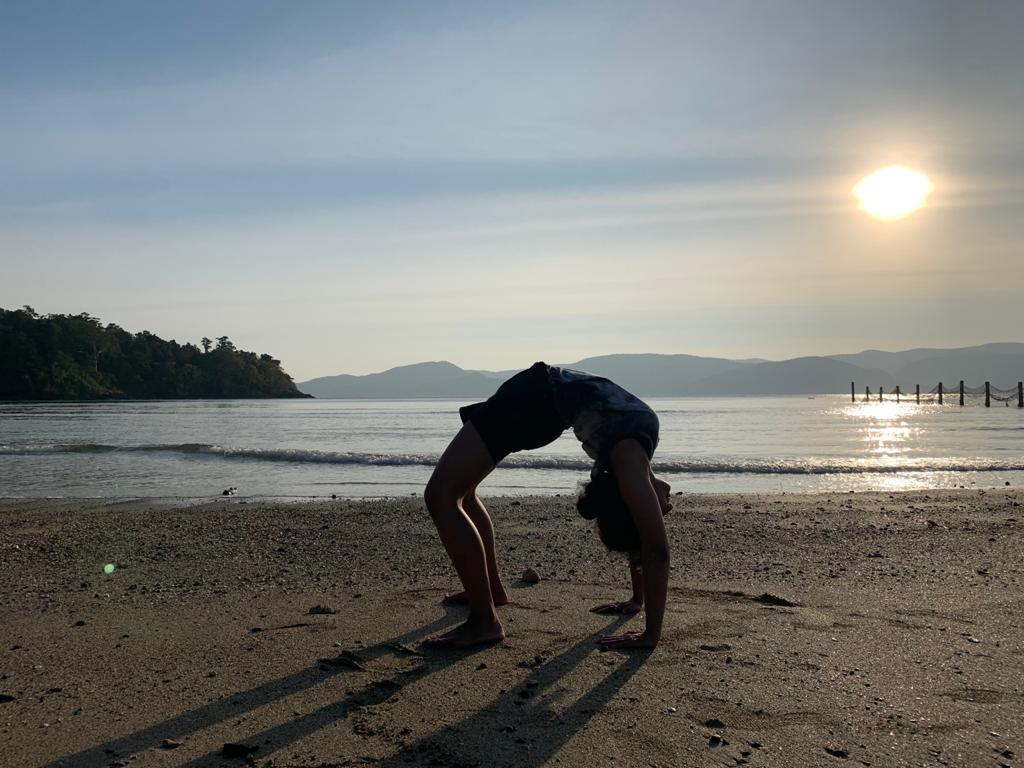 Person performing a backbend on a beach with water, trees, and a setting sun in the background.