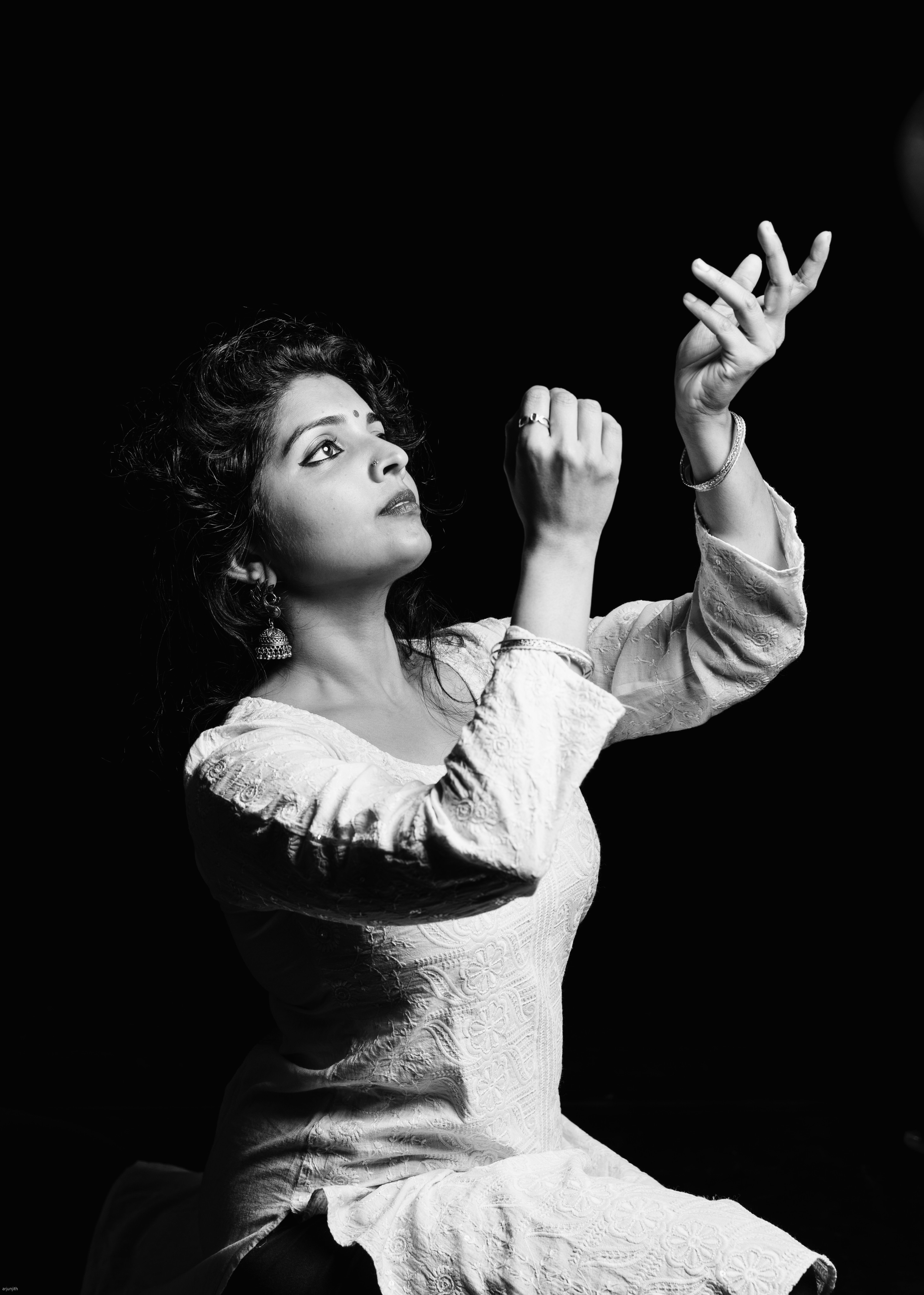 Woman with curly hair looking upward, raising her right hand, wearing a light-colored dress, against a dark background.