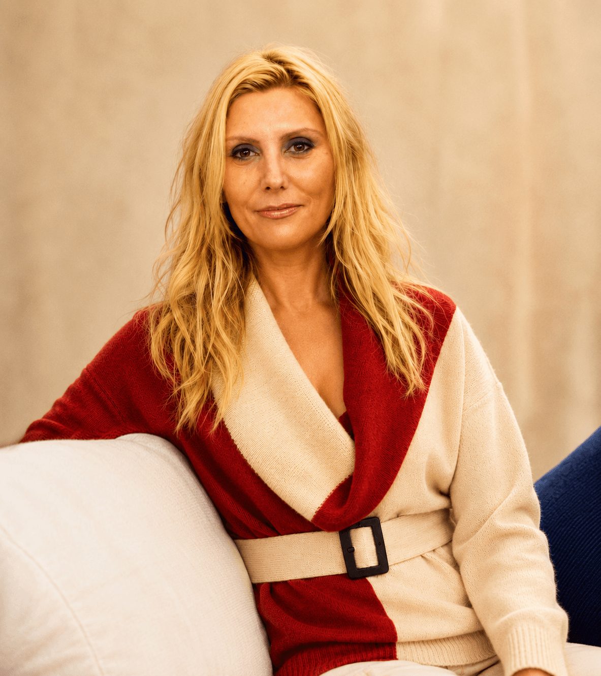 Woman with blonde hair wearing a red and beige sweater, sitting on a white couch, neutral background.