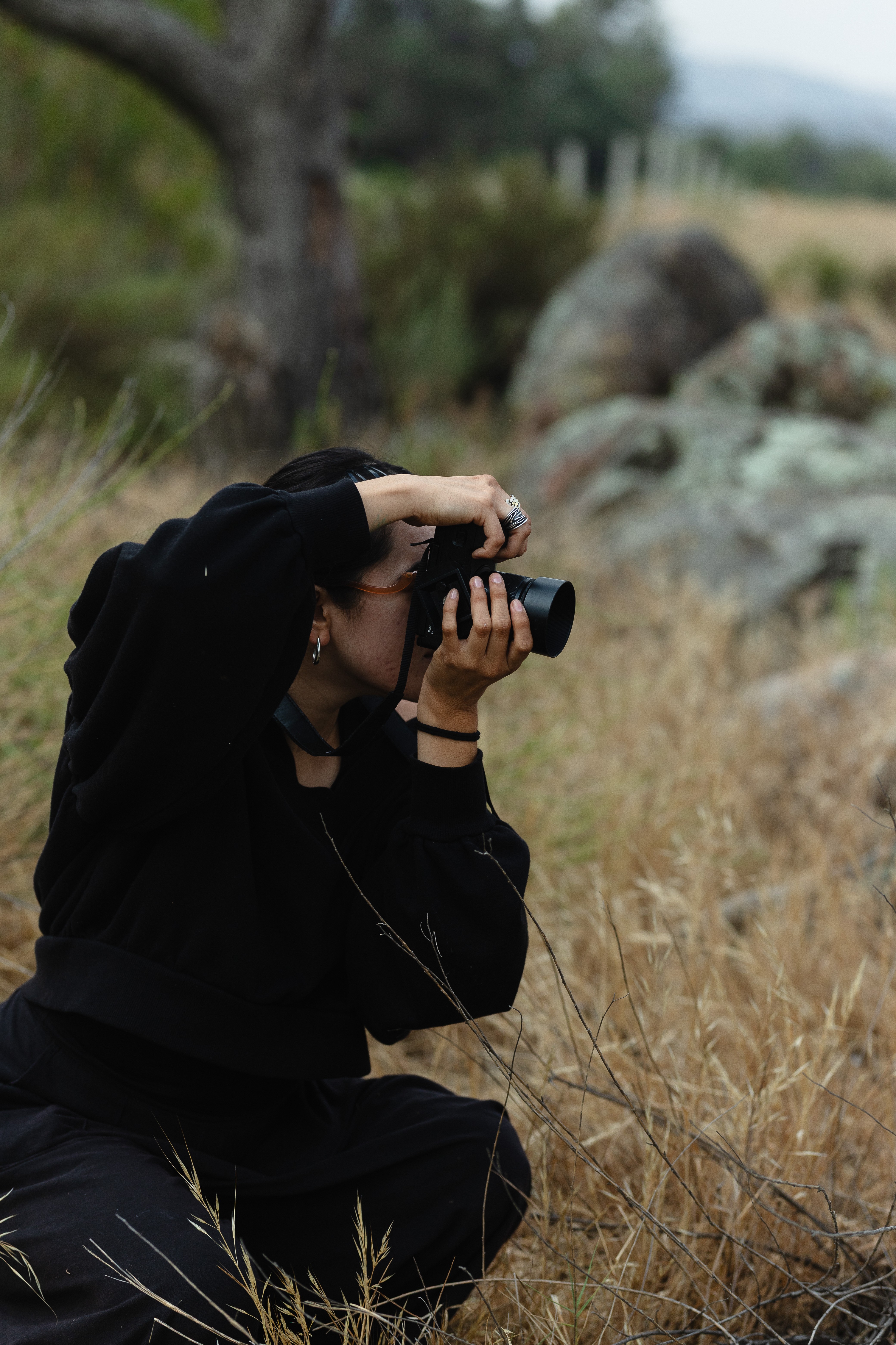 Person crouching outdoors, holding a camera to their eye, surrounded by grass and rocks, with trees in the background.