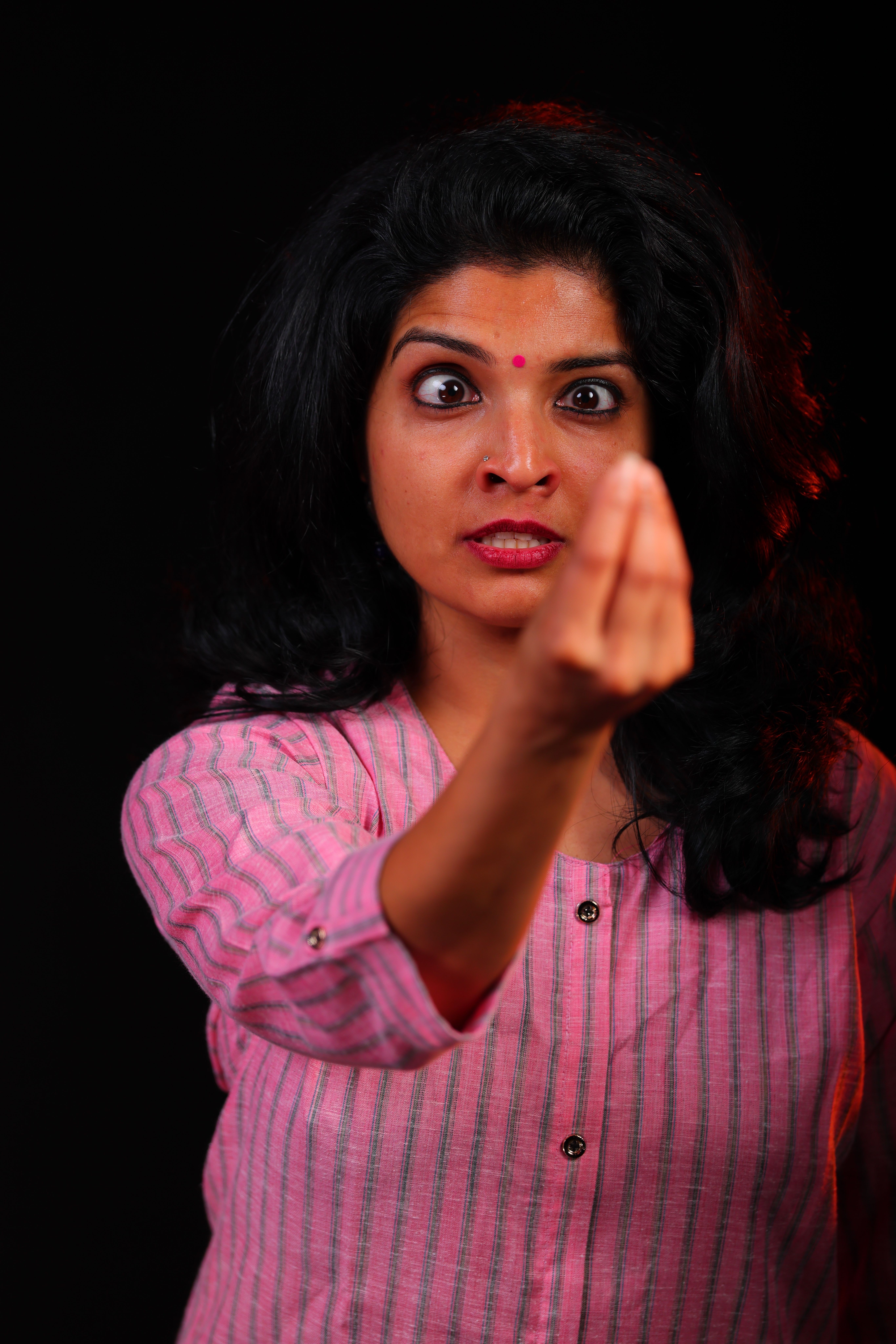 Woman with long black hair wearing pink striped shirt, covering her mouth with her hand, looking at the camera.
