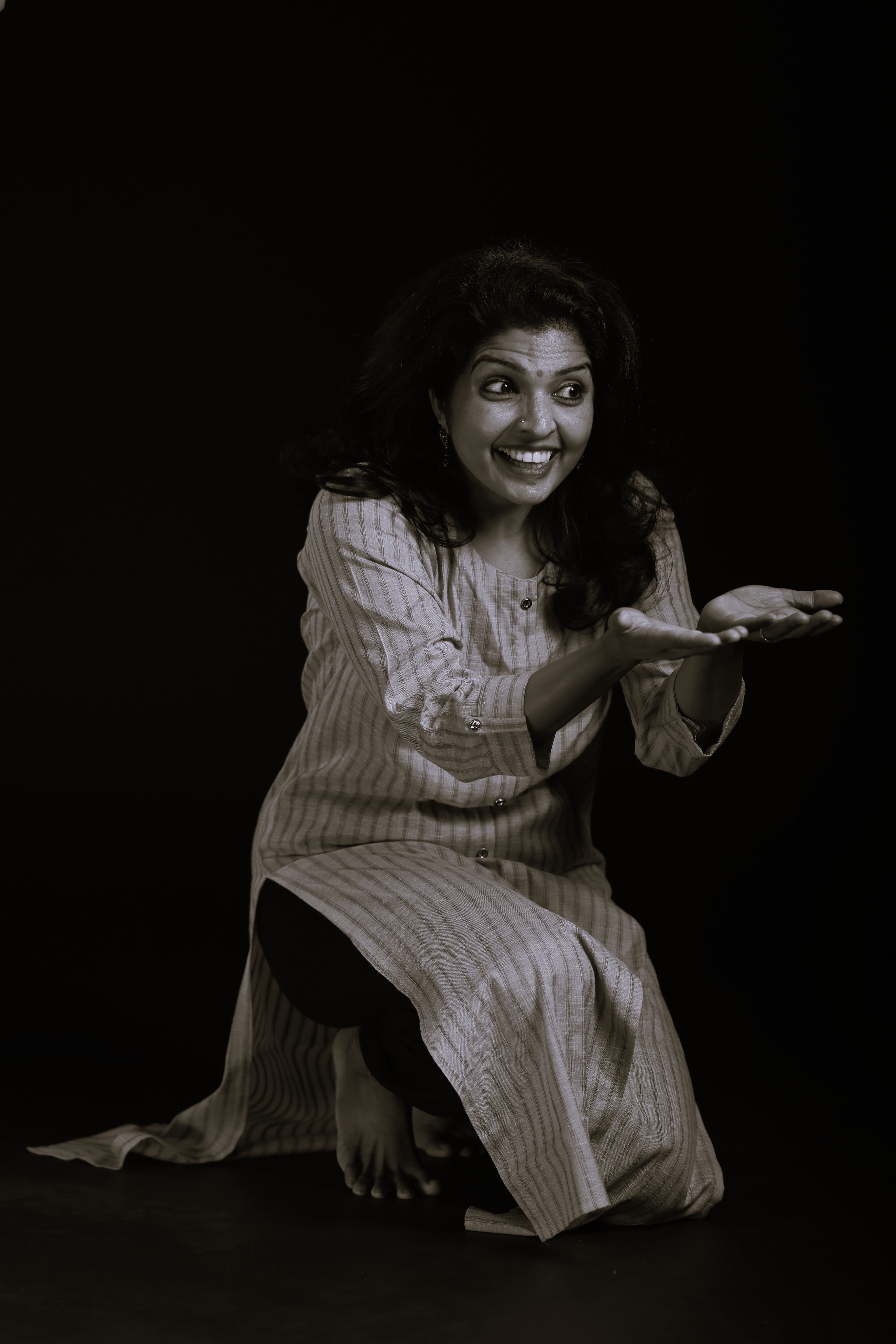 Woman crouching with hands extended, smiling, wearing a striped dress, against a dark background.