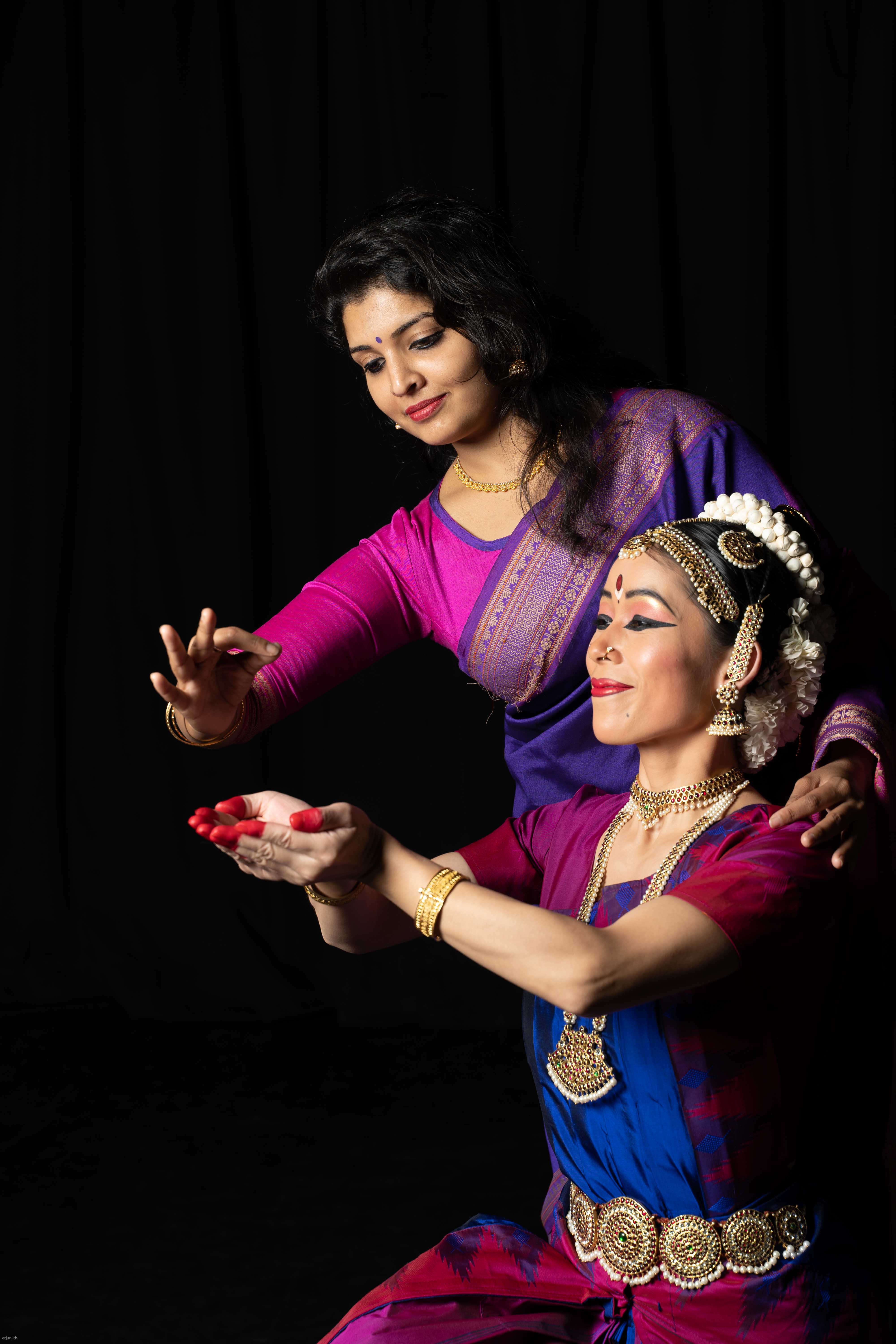 Two women in traditional attire, one performing a dance with hands raised, the other observing, against a black background.