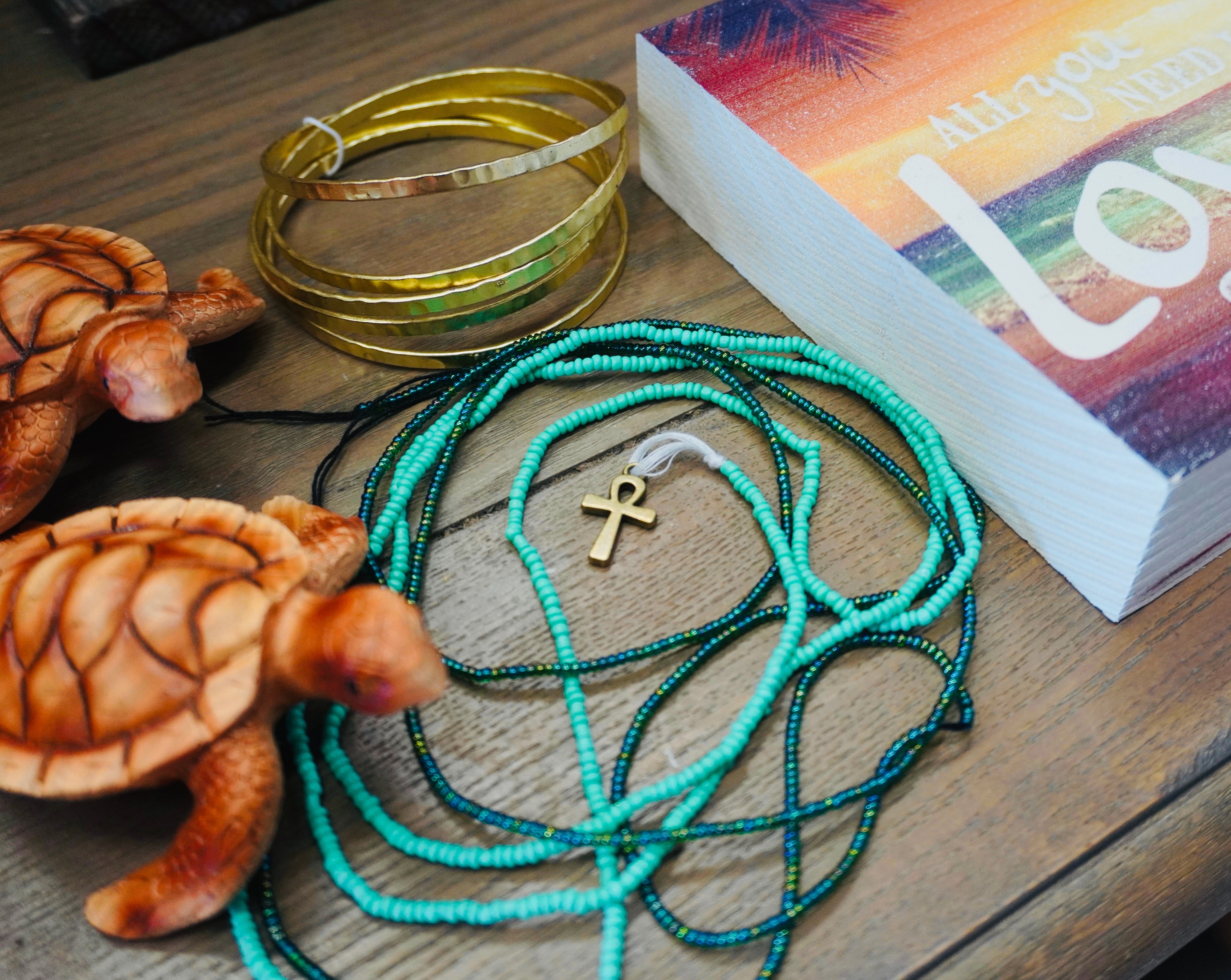 Turquoise and gold jewelry, a gold cross necklace, a book with a colorful cover, and two turtle figurines on a wooden surface.