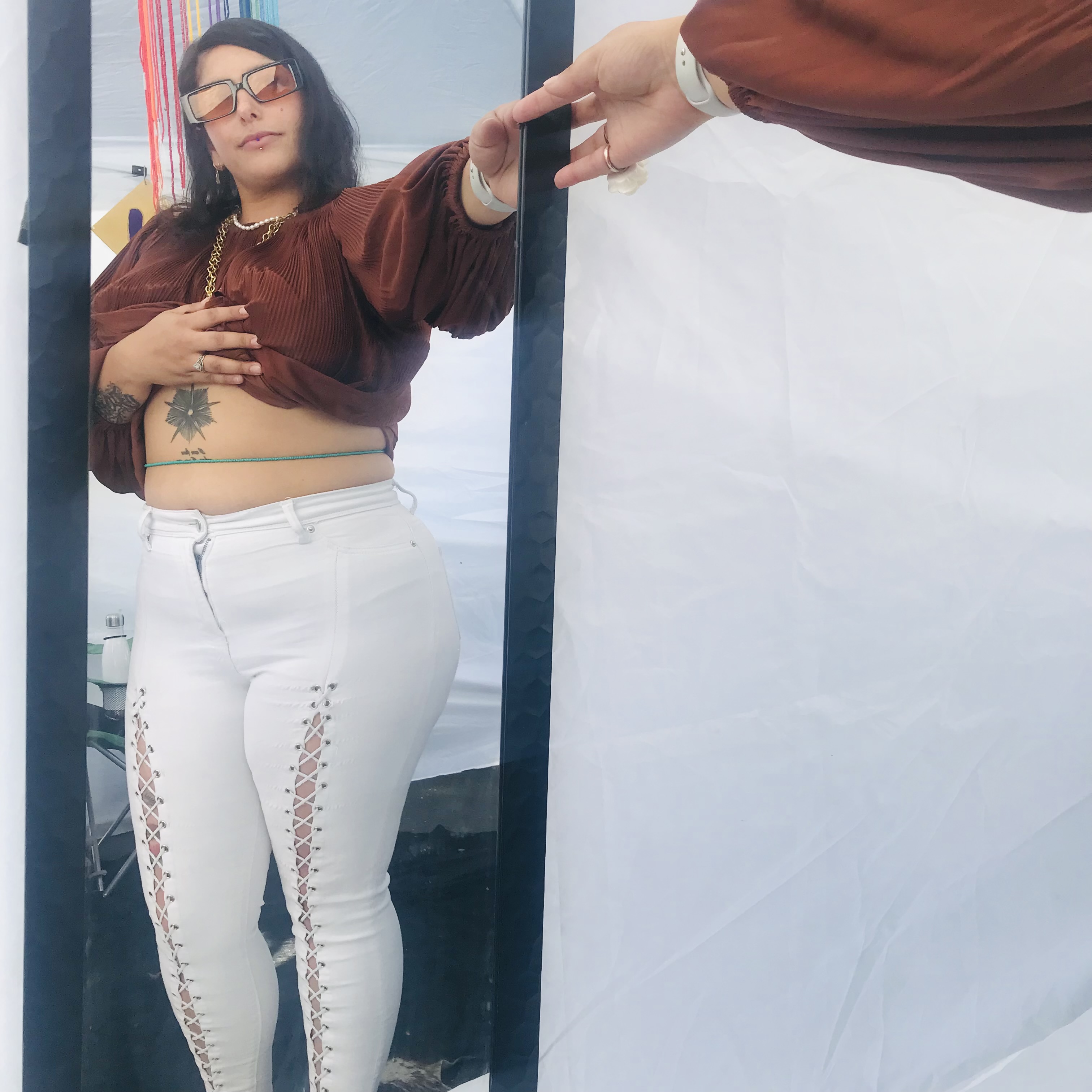 Person taking a mirror selfie outdoors, wearing glasses, a brown top, and white pants with lace-up details, holding a phone.