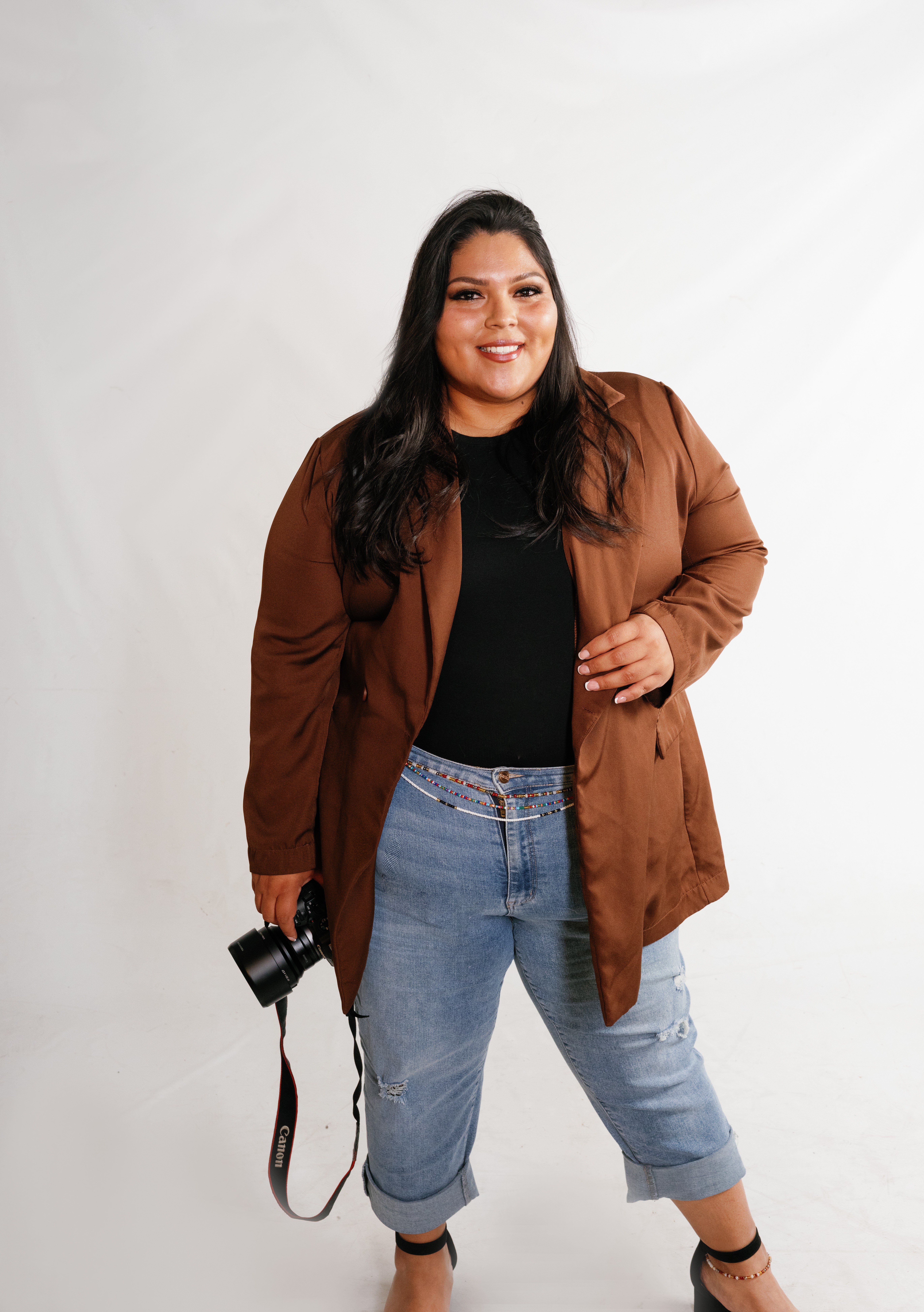 Woman with long dark hair smiling, wearing a brown jacket, black top, and jeans, holding a camera, standing against a white background.
