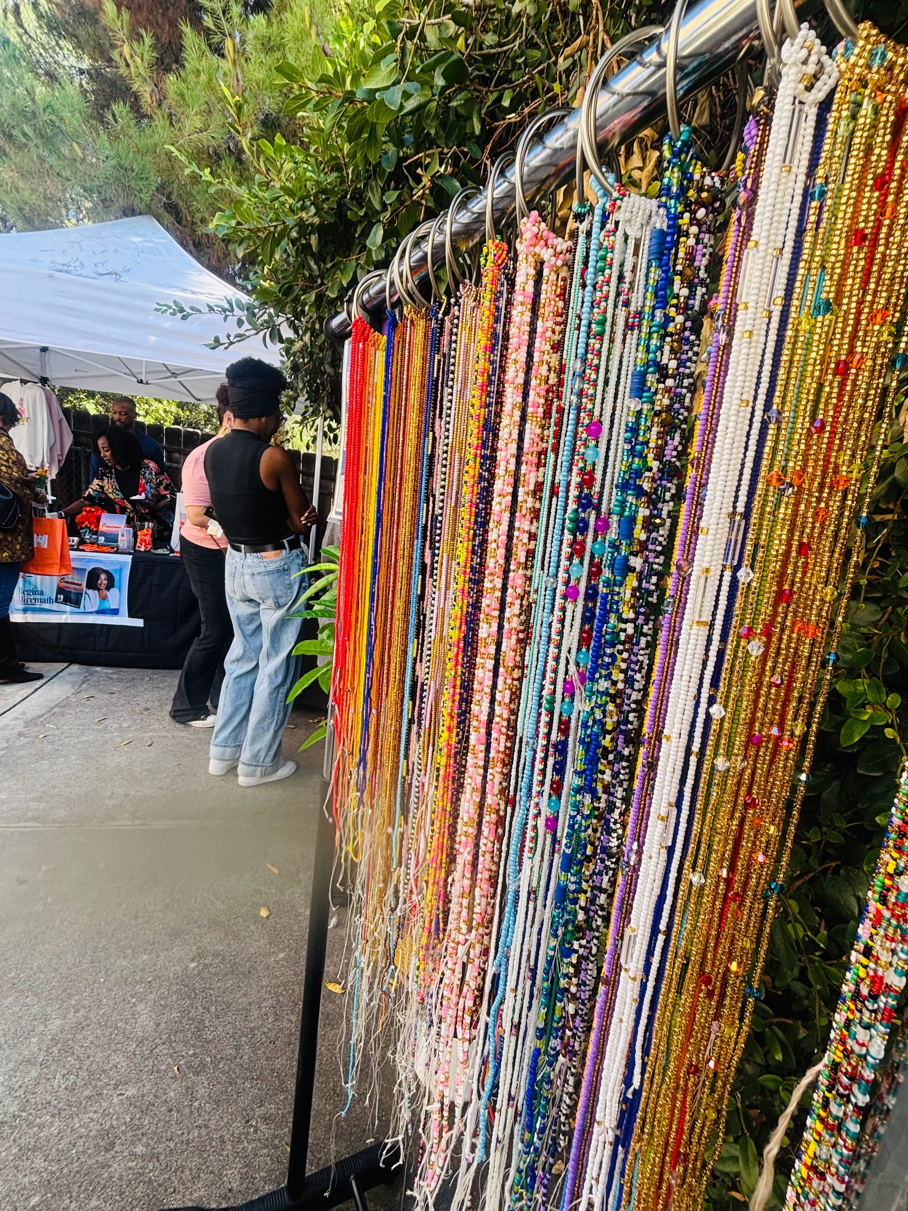 Colorful beaded necklaces hanging on a display rack at an outdoor market, with people and a tent in the background.